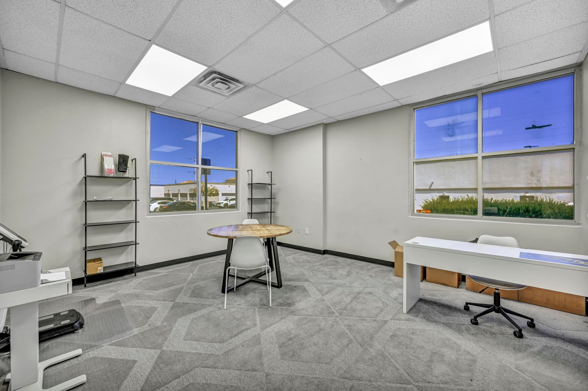 Office interior with gray patterned carpet, large windows, a round table, and a white desk.