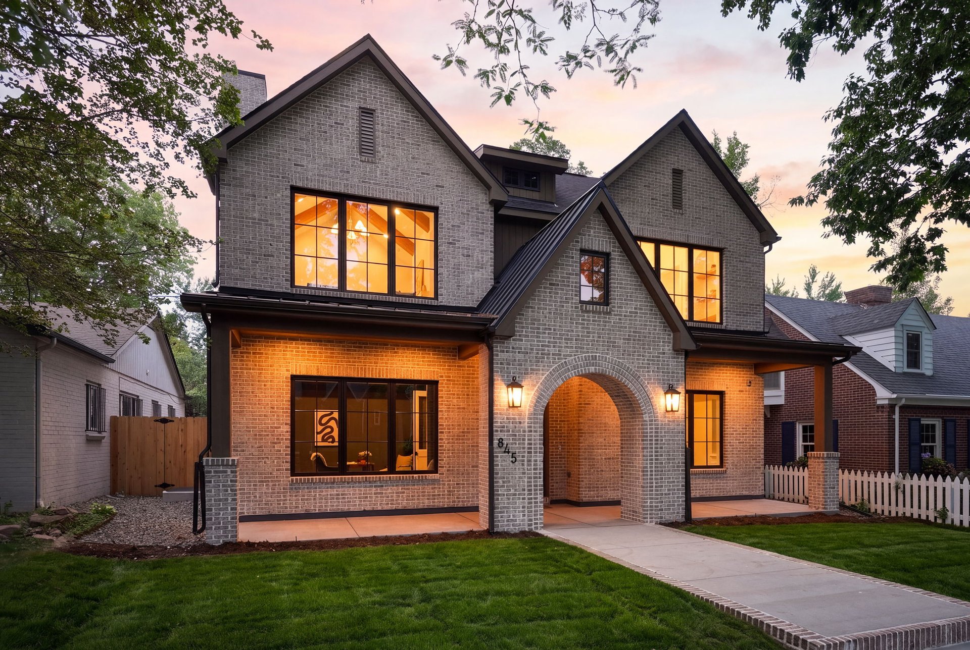 Two-story brick house with a gray arched entry and warmly lit windows at dusk.