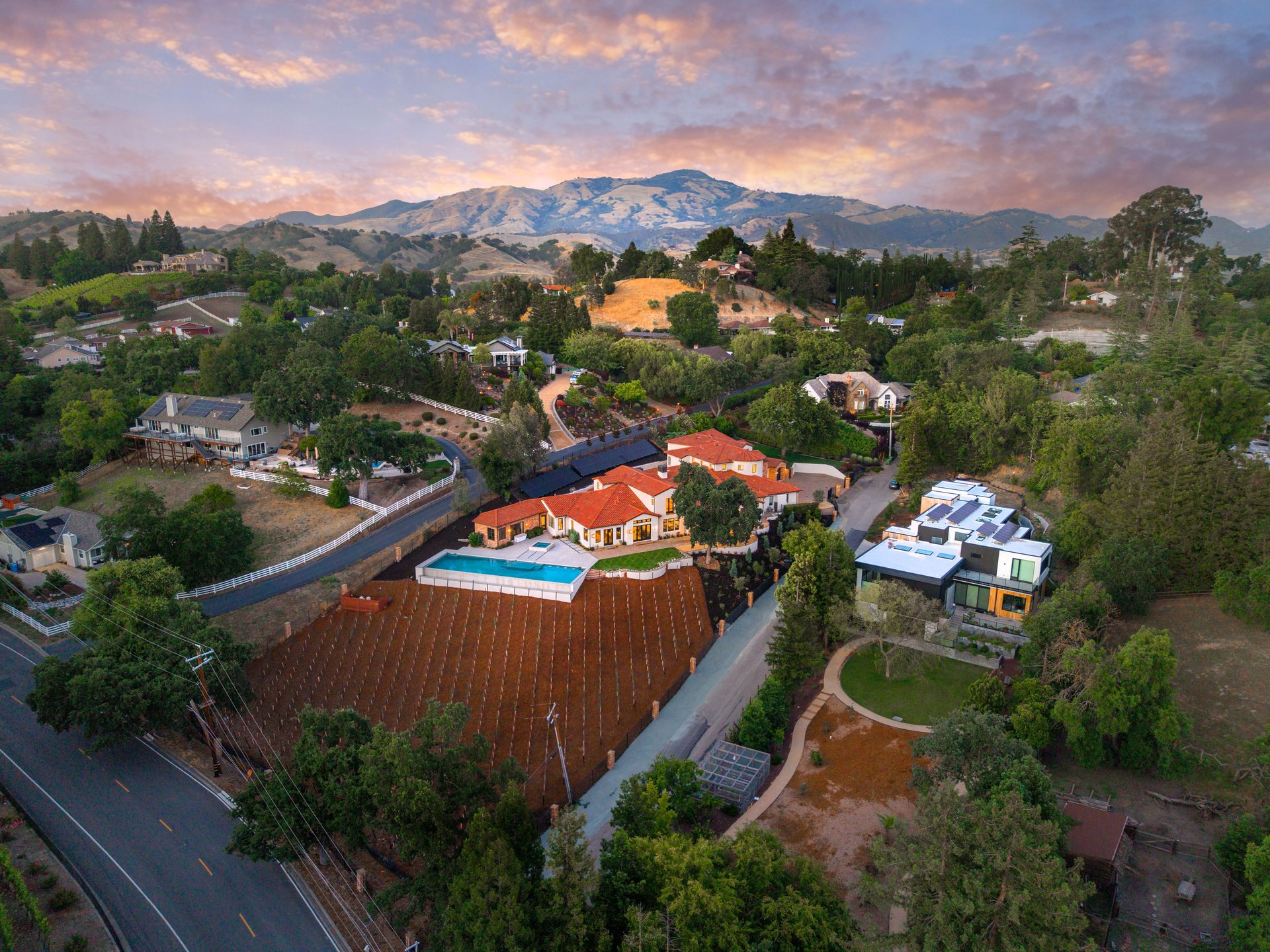 Aerial view of upscale hillside homes, a pool, vineyards, and mountains at sunset.