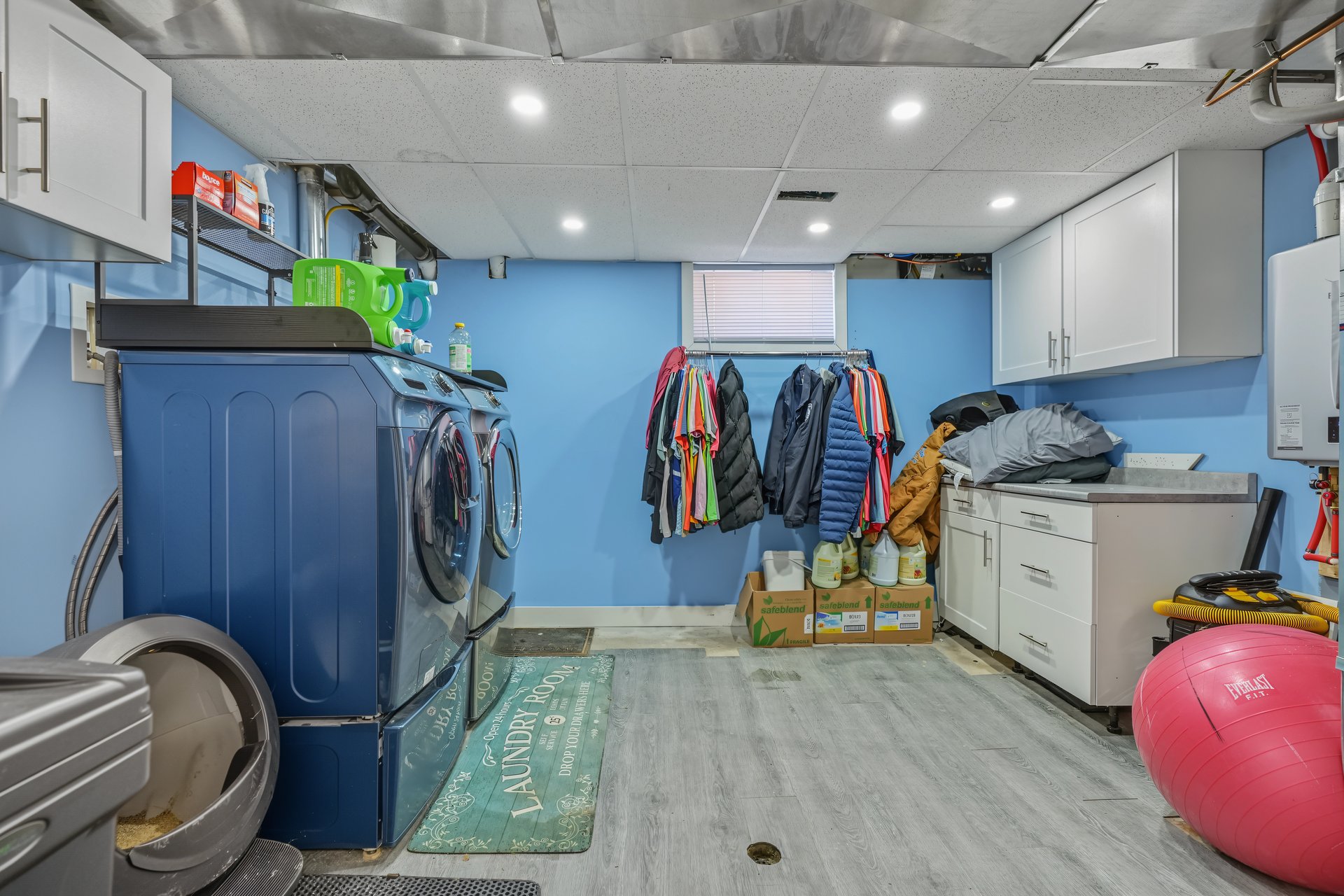 Laundry room with multiple machines, white cabinets, and clothes hanging beneath a window.