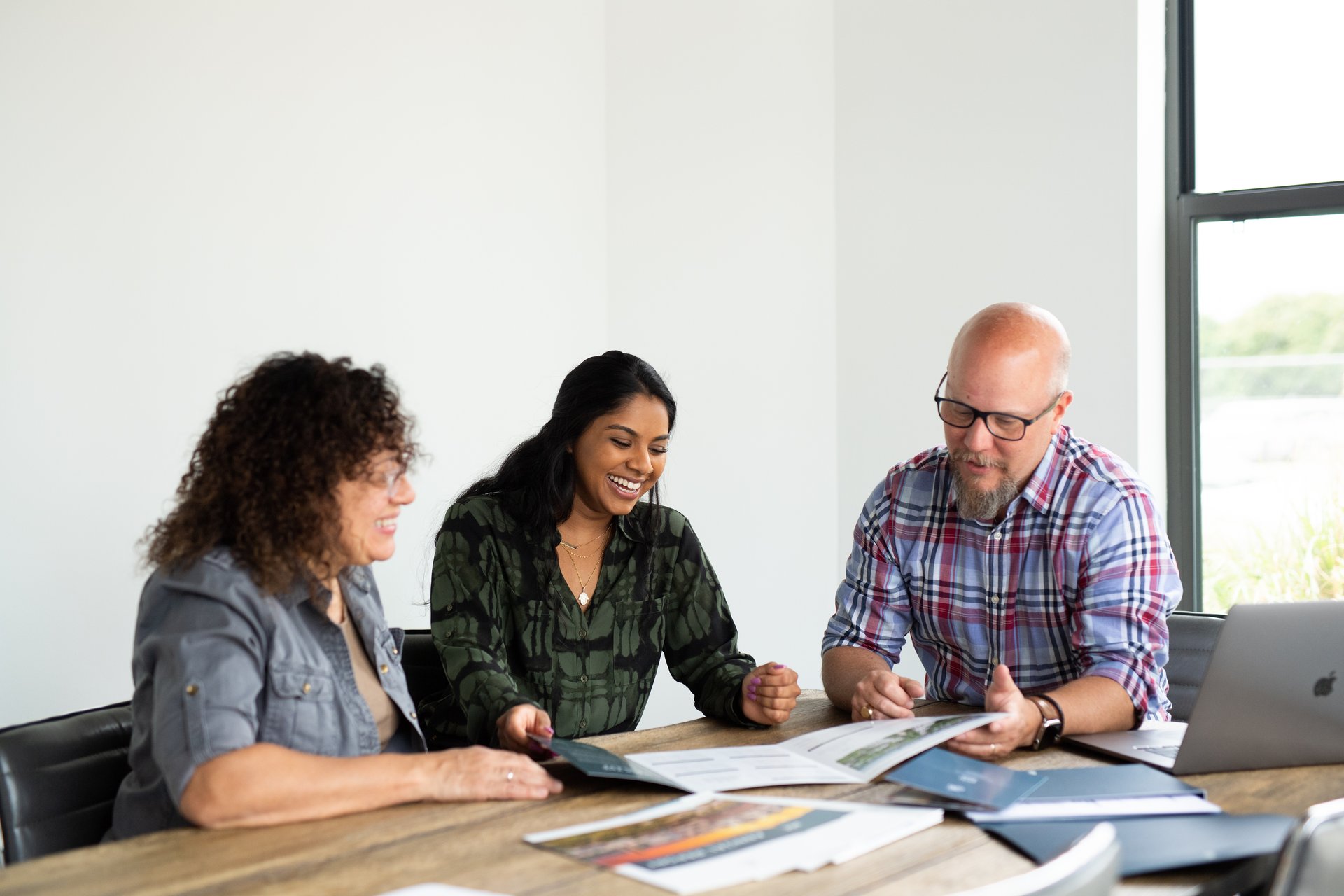 Three adults review printed materials and a laptop together at a bright office table.