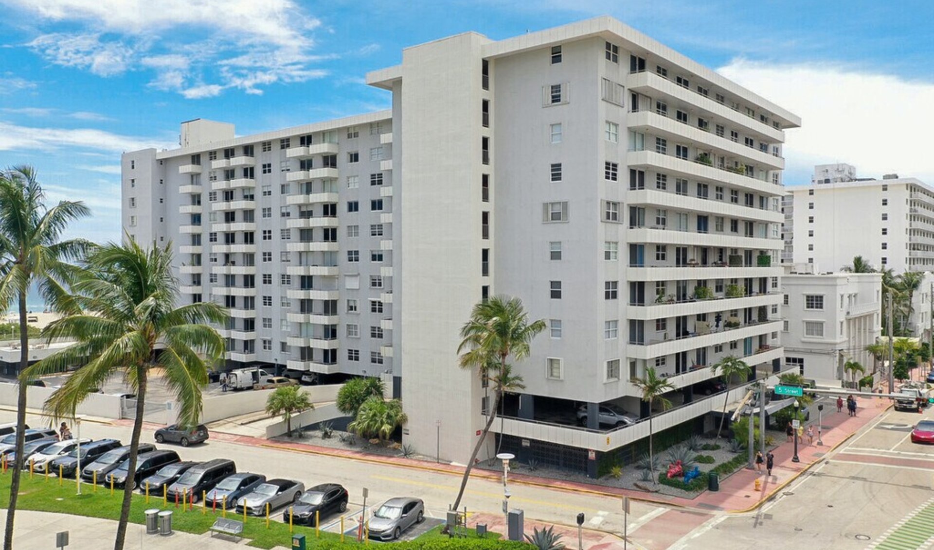 White beachfront condominium buildings with long balconies, palm trees, and a street-level parking area.