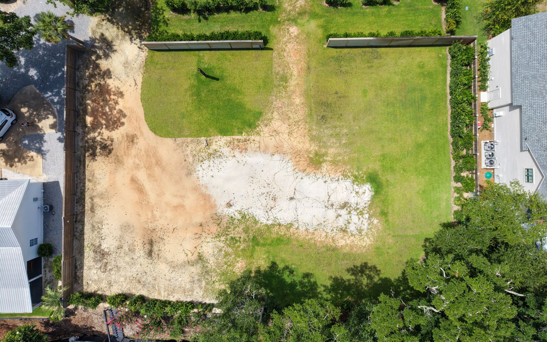 Vacant lot with a grassy lawn and sandy area, viewed from above beside neighboring homes.