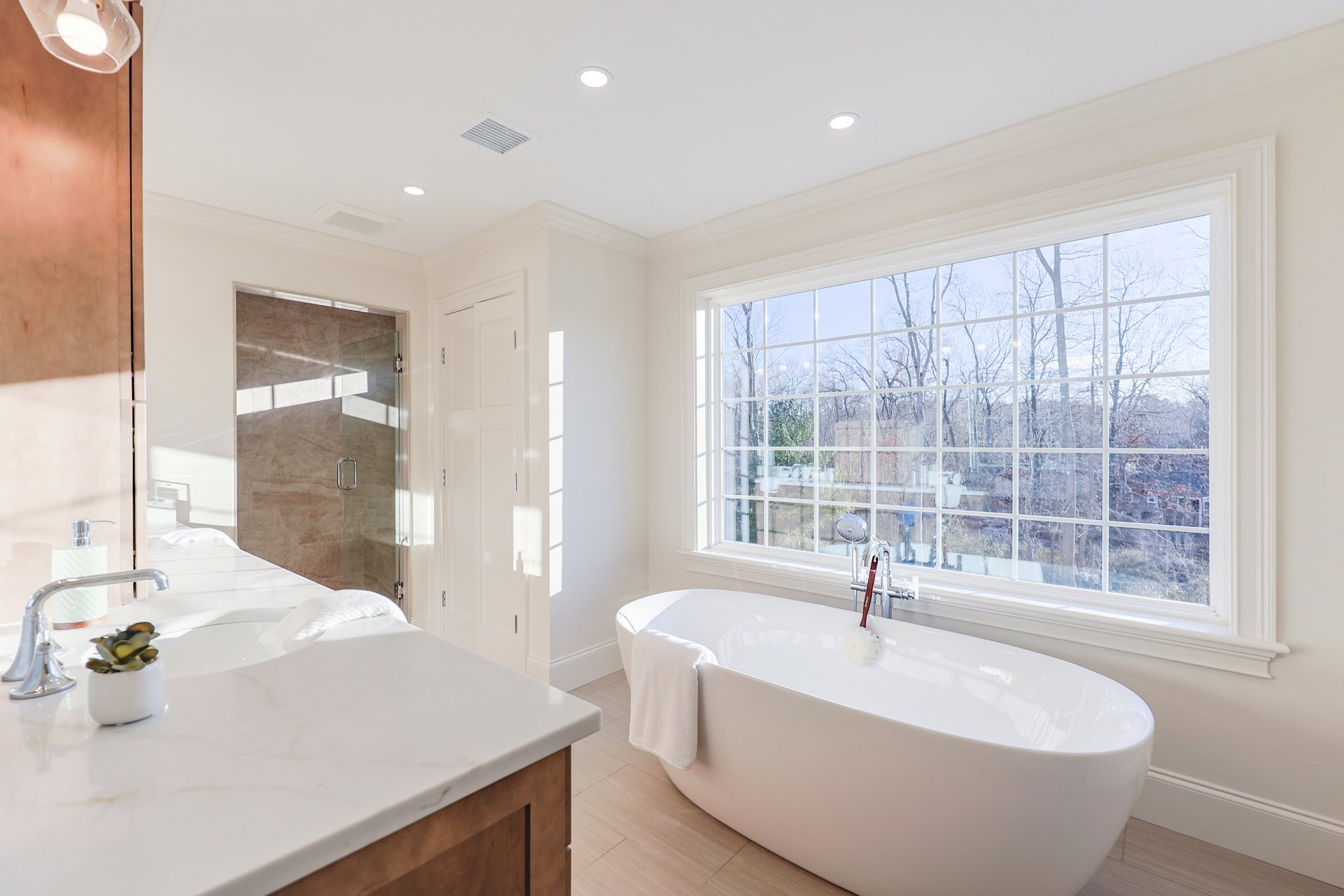 Bathroom with a freestanding tub beside a large window, marble vanity, and glass-enclosed shower.