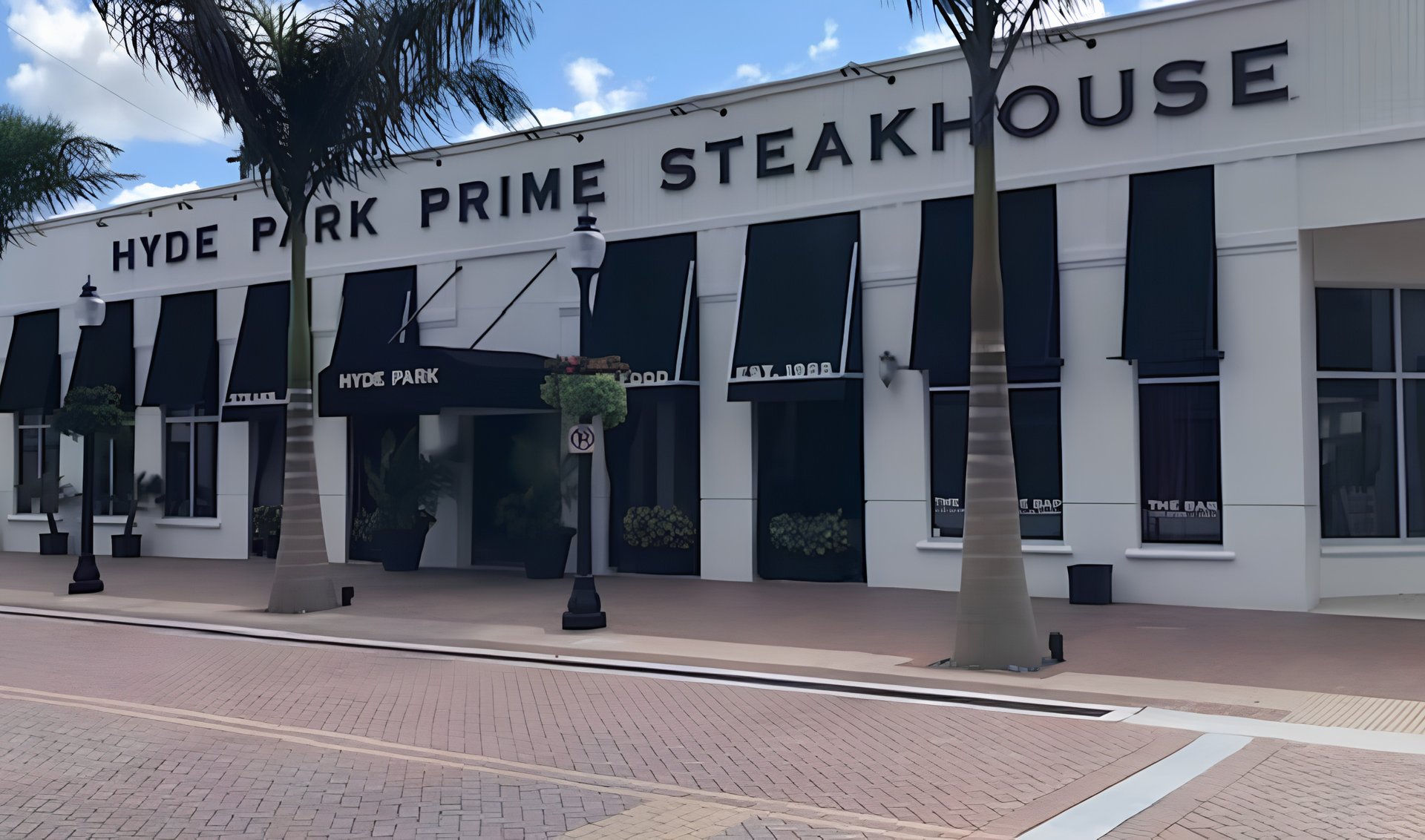 Hyde Park Prime Steakhouse exterior with palm trees and a brick-paved sidewalk.
