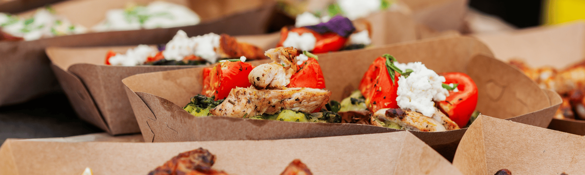 A close-up of several food trays featuring grilled chicken, roasted tomatoes, greens, and toppings like cheese and herbs.