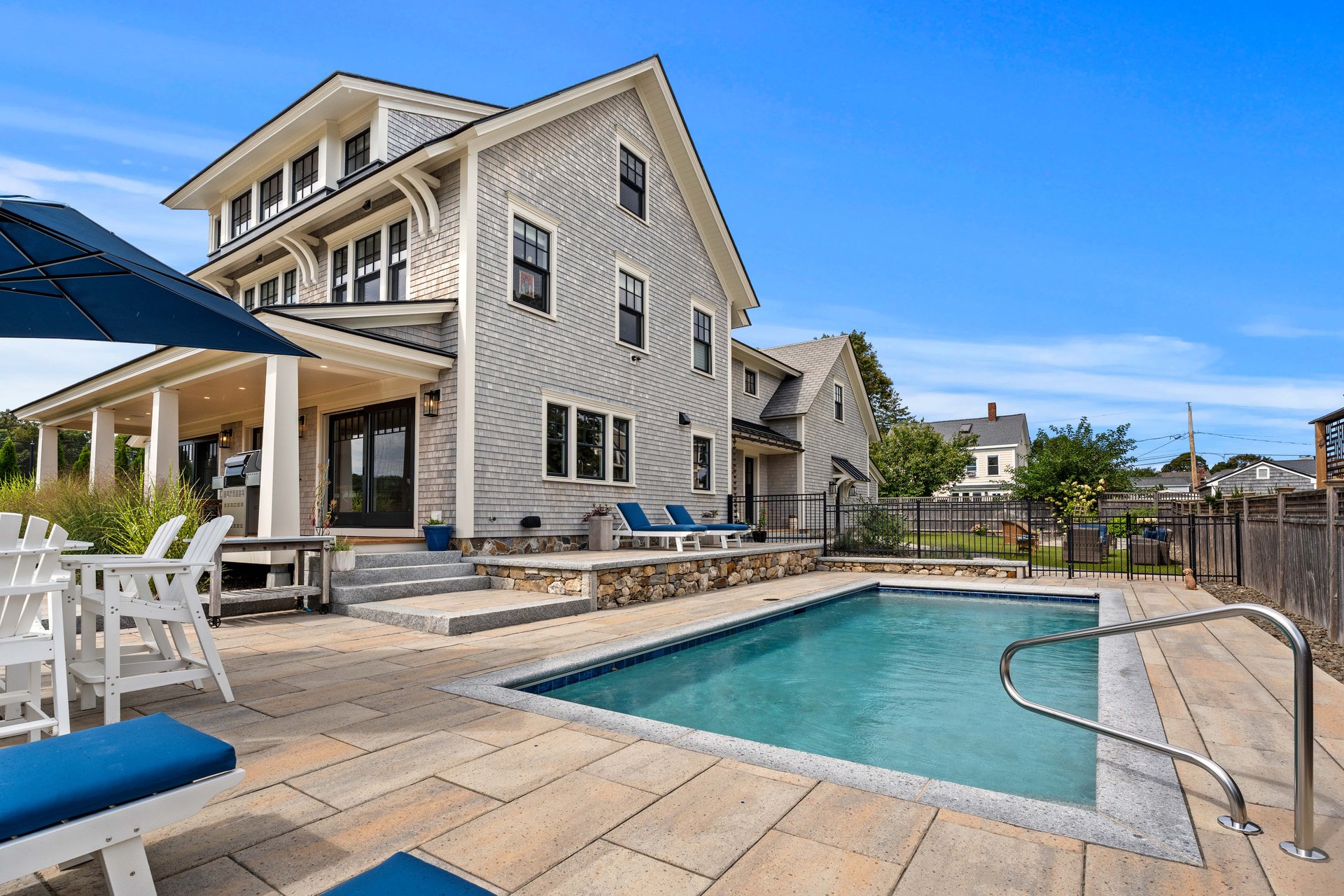 Gray-shingle home beside a swimming pool, with a broad stone patio and outdoor seating.