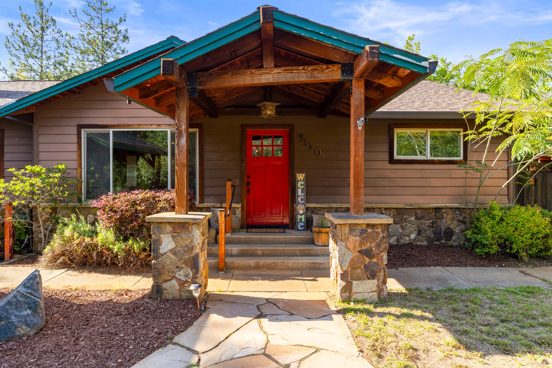 Home entrance with a red front door beneath a timber porch and teal-trimmed roof.