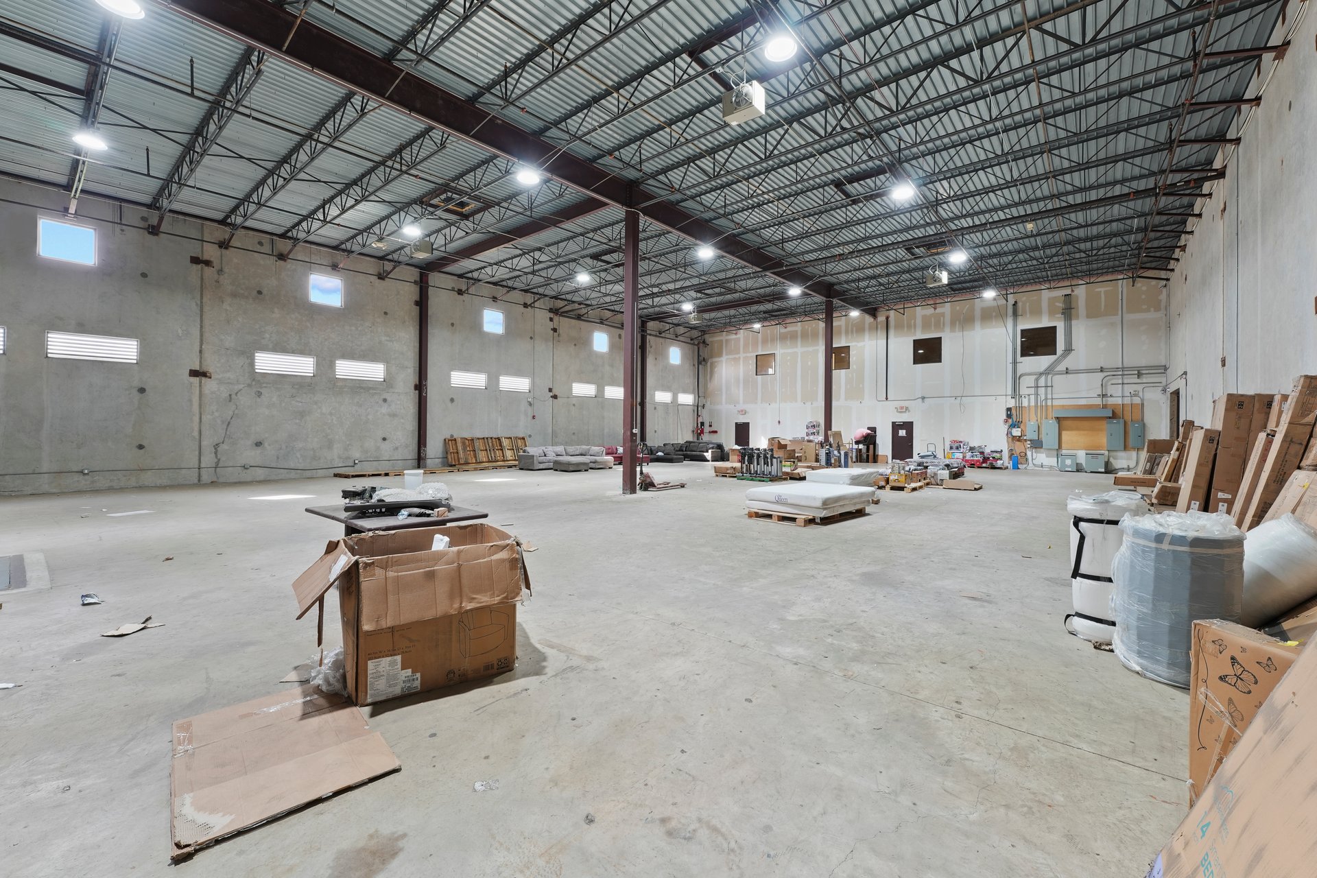 Large warehouse interior with exposed steel roof trusses, concrete floor, and scattered boxes and equipment.