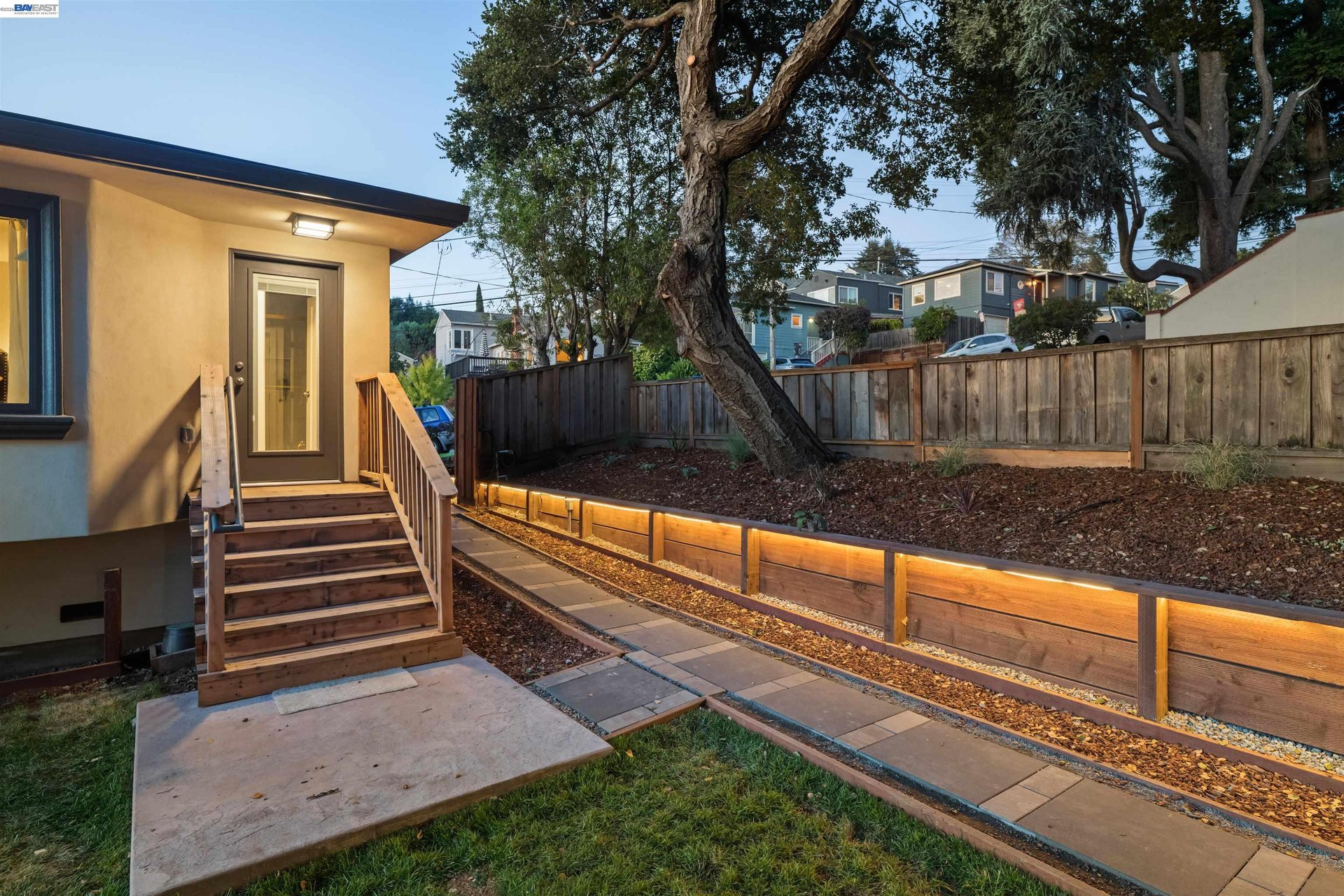 Backyard with wooden entry steps, paved walkway, raised garden beds, and a large mature tree.