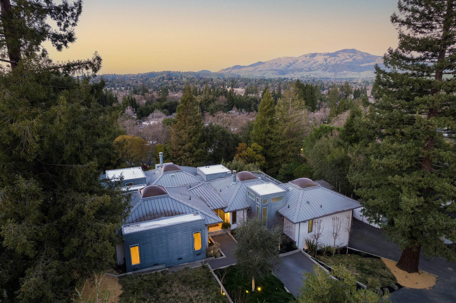 Aerial view of a modern home among trees, with a wooded neighborhood and mountains beyond.