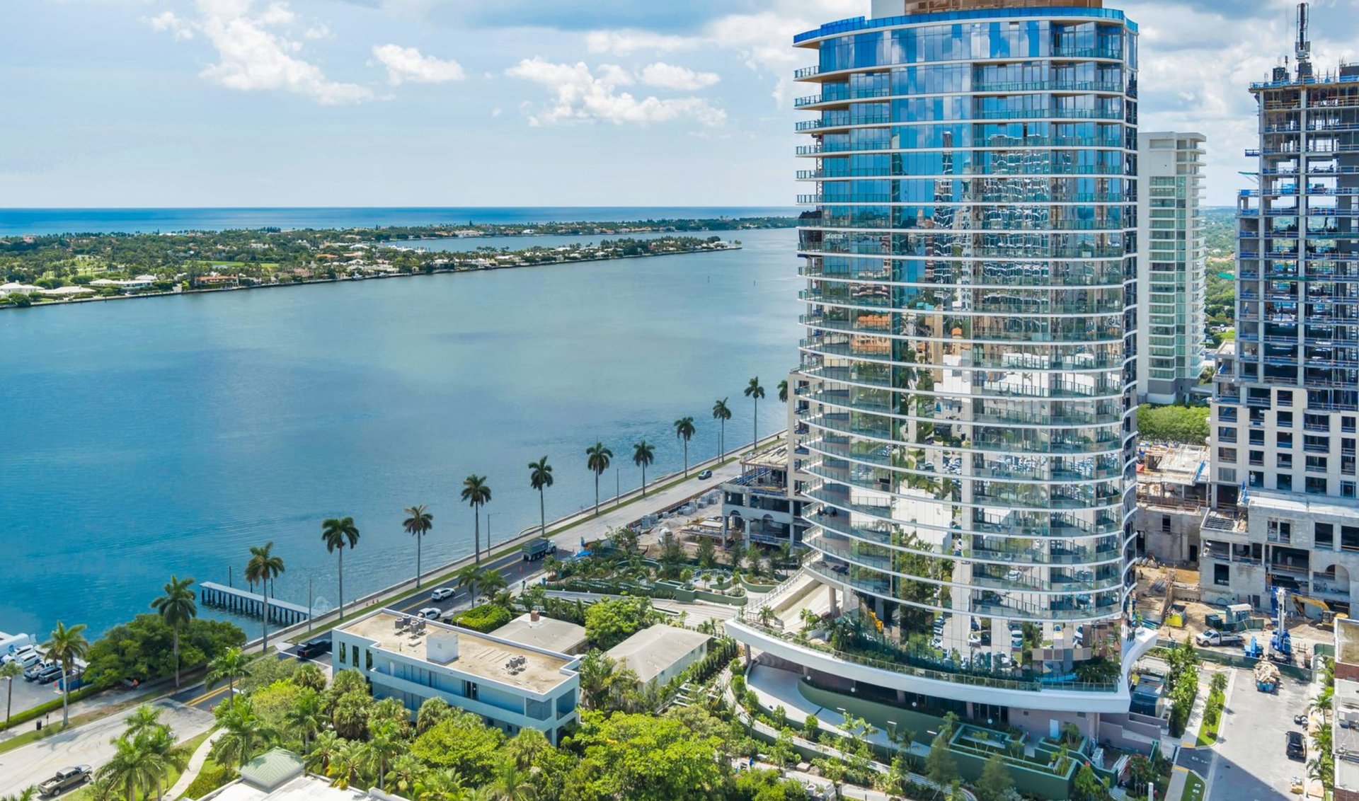 Glass condominium tower beside the waterfront, with palm-lined roads and a neighboring tower under construction.