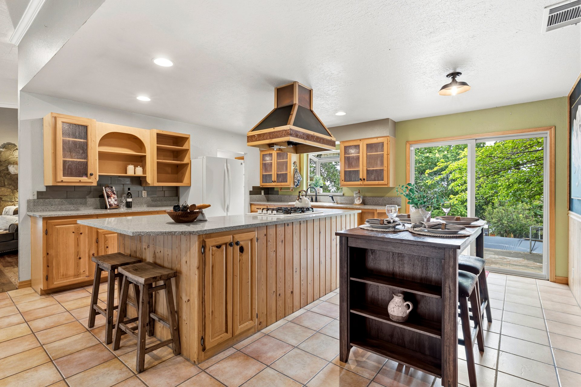 Kitchen with natural wood cabinetry, a stone-counter peninsula, and a dining island beside a leafy view.