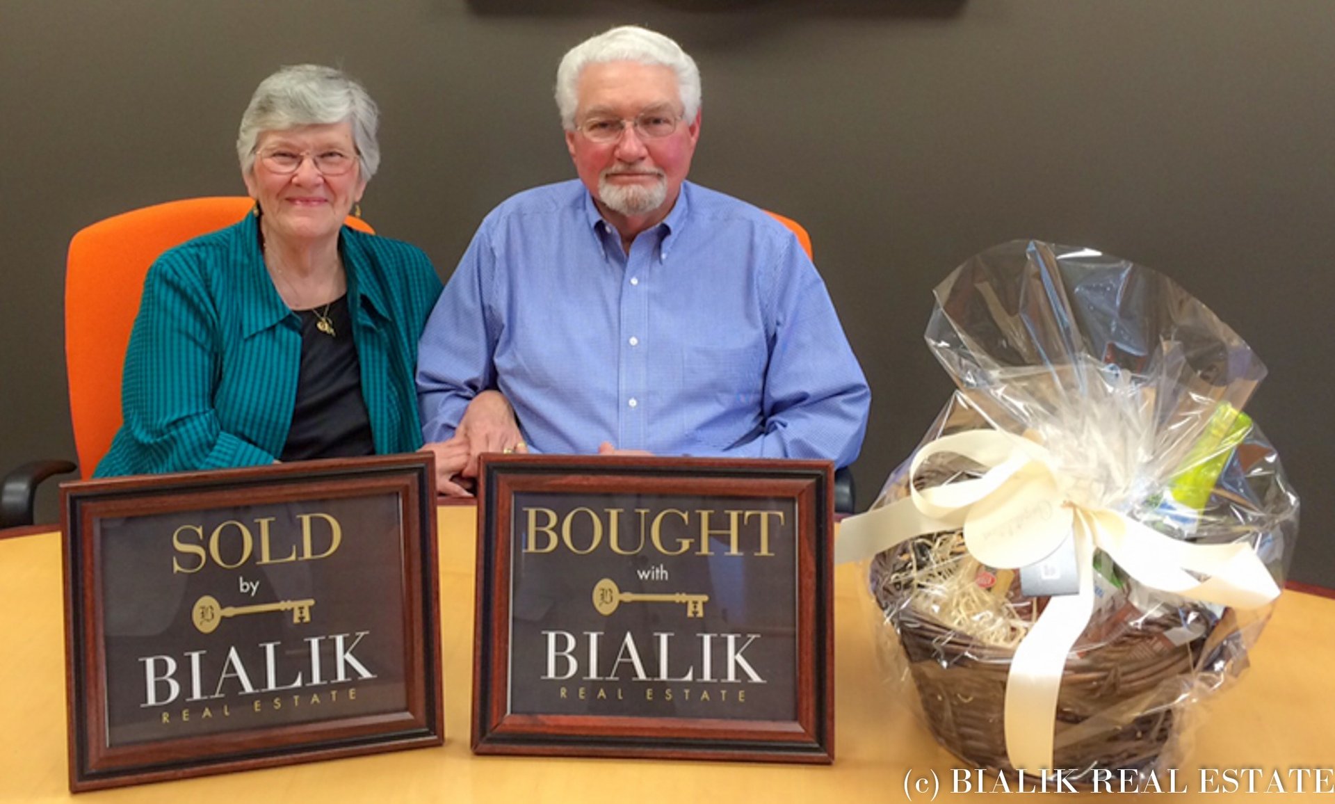 Older couple seated behind framed SOLD and BOUGHT signs, beside a wrapped gift basket.