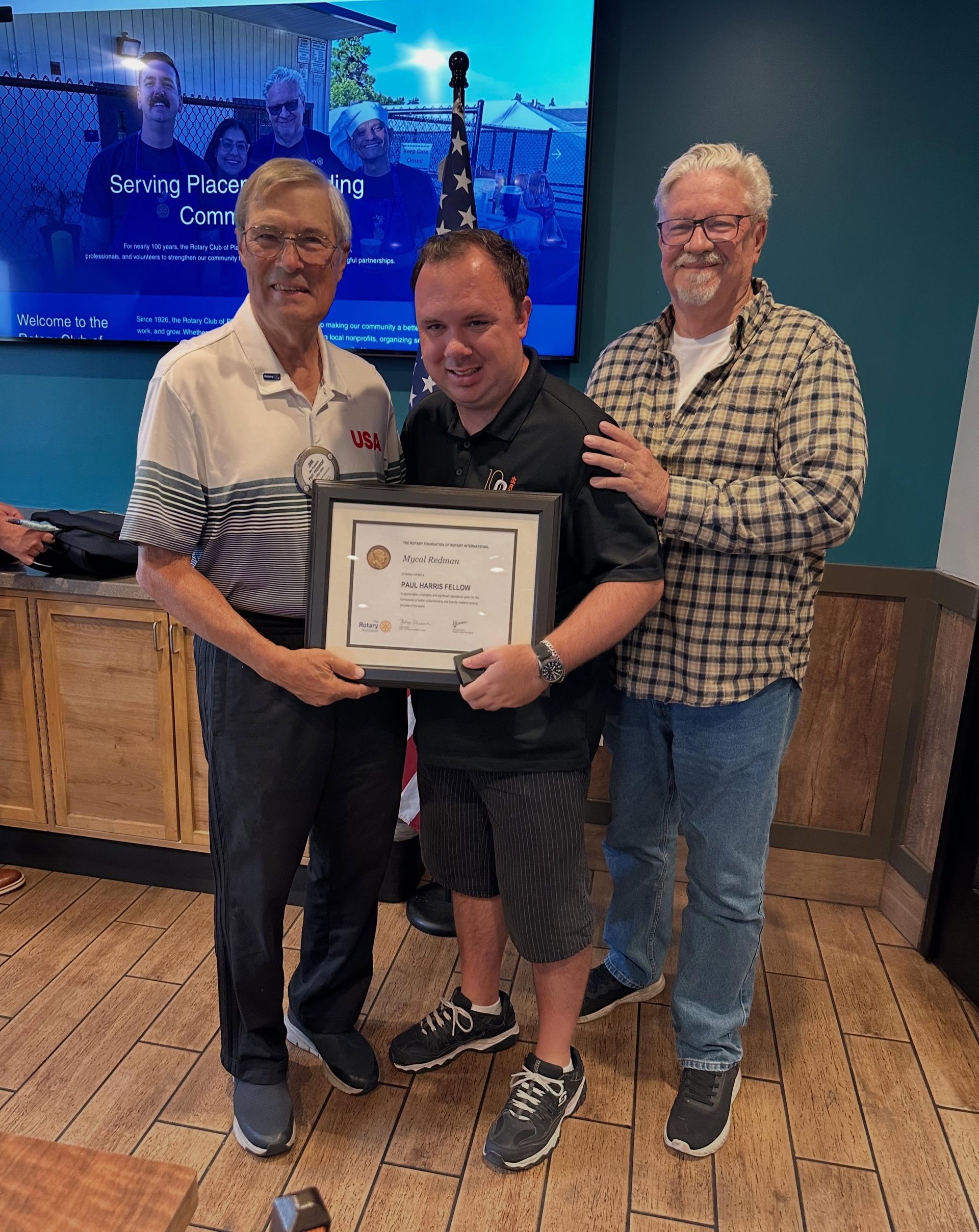 Three men pose with a framed Paul Harris Fellow certificate beside an American flag.