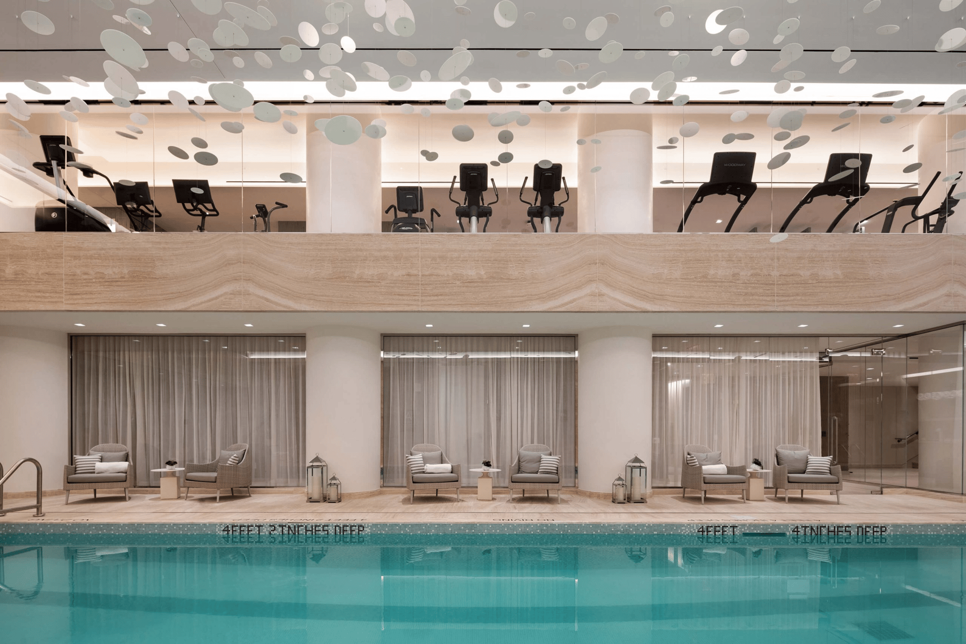 Indoor pool with lounge seating and an upper-level fitness center beneath suspended ceiling decorations.