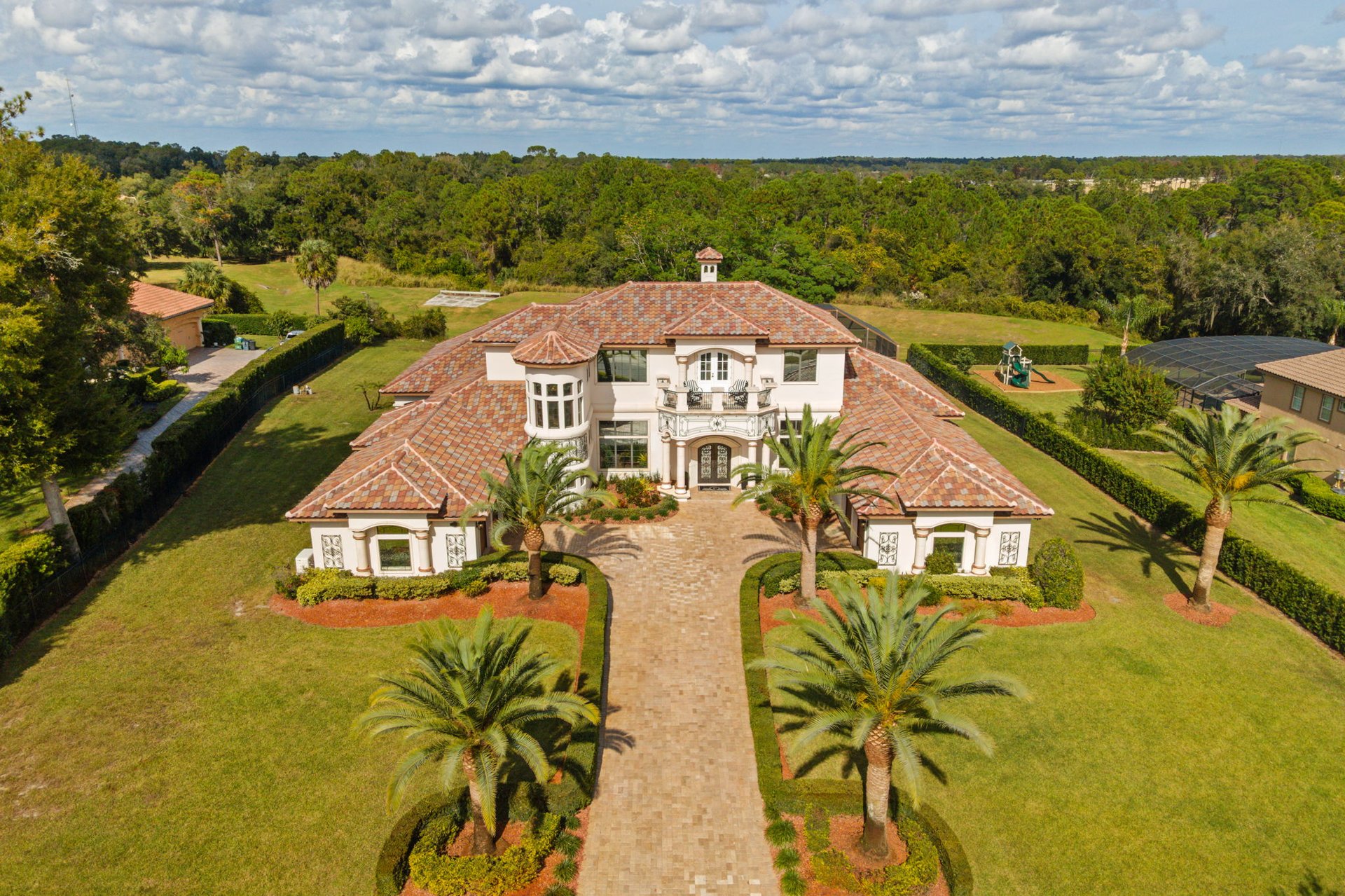 Aerial view of a large tiled-roof home with a palm-lined walkway and landscaped grounds.