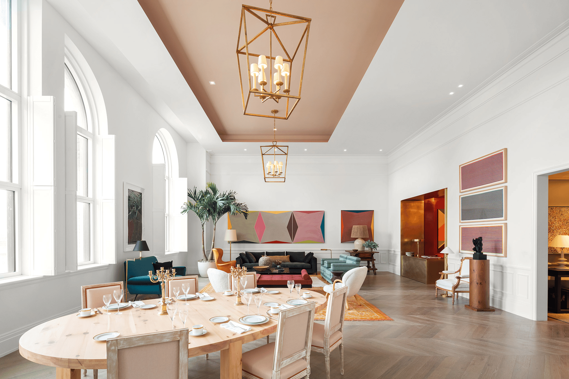 Furnished communal dining room with arched windows, a set table, colorful artwork, and brass chandeliers.