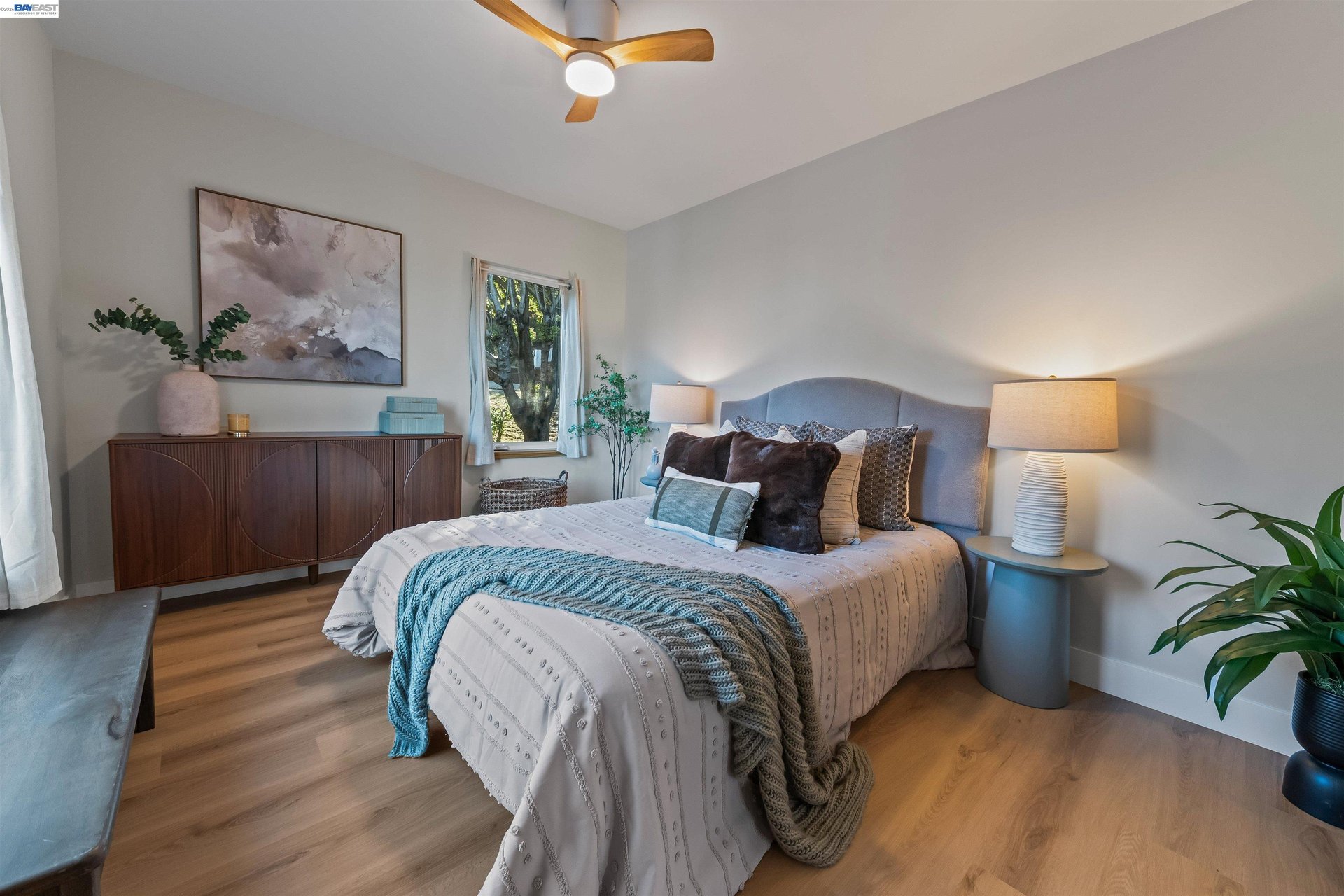 Staged bedroom with a large bed, blue knitted throw, wood sideboard, and ceiling fan.