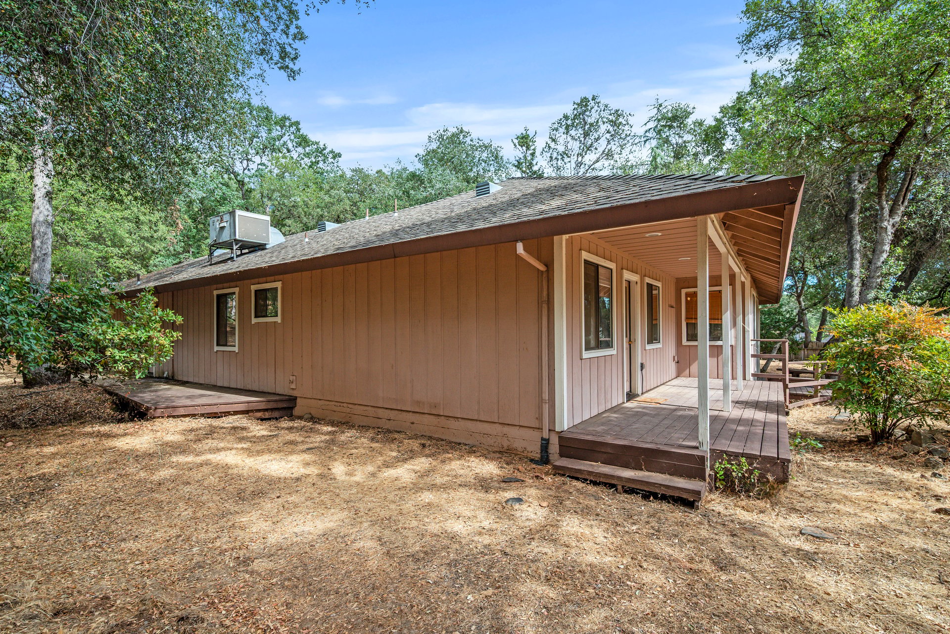 Single-story brown home with a covered porch and raised deck among trees.