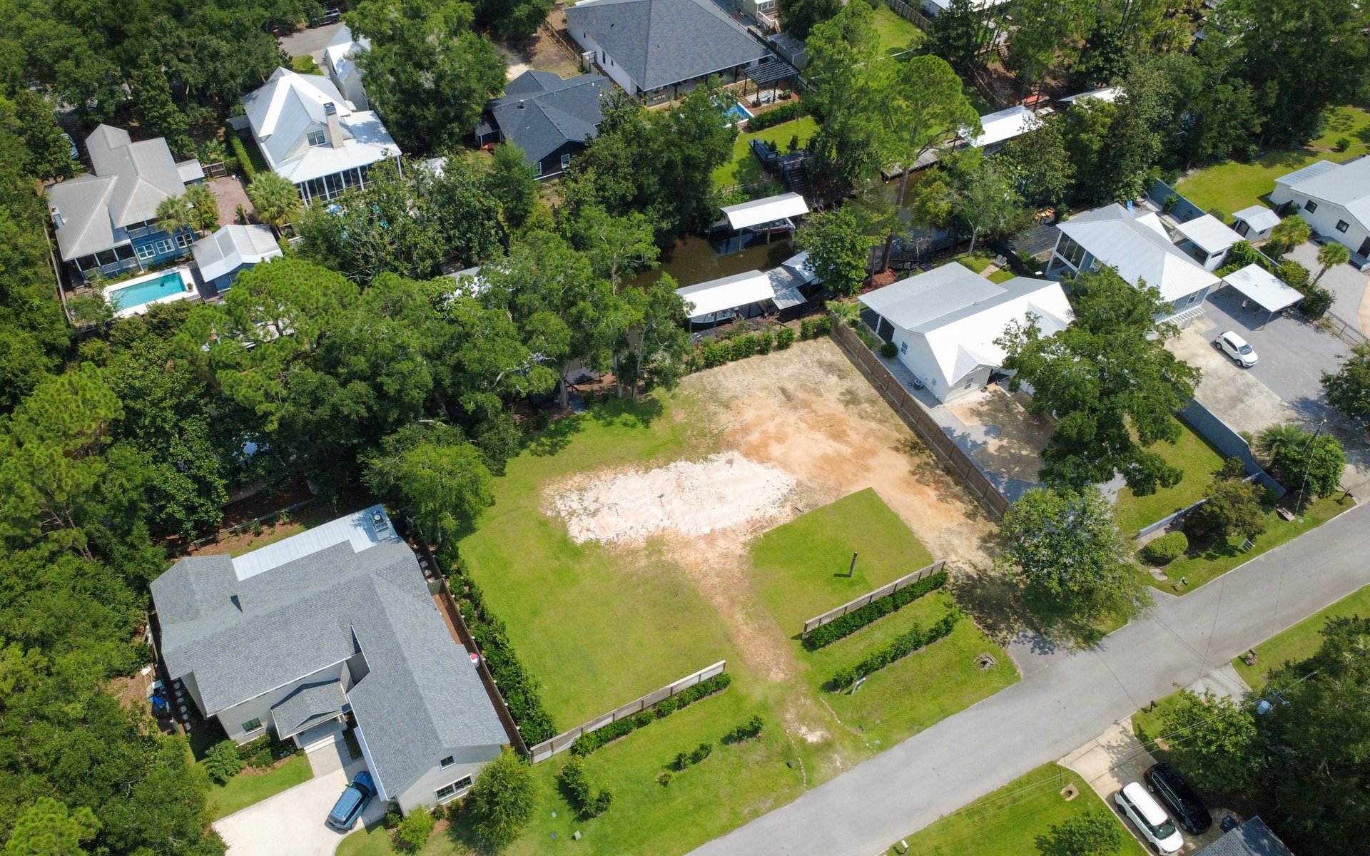 Cleared vacant lot surrounded by homes and trees, with a canal visible behind the neighboring properties.
