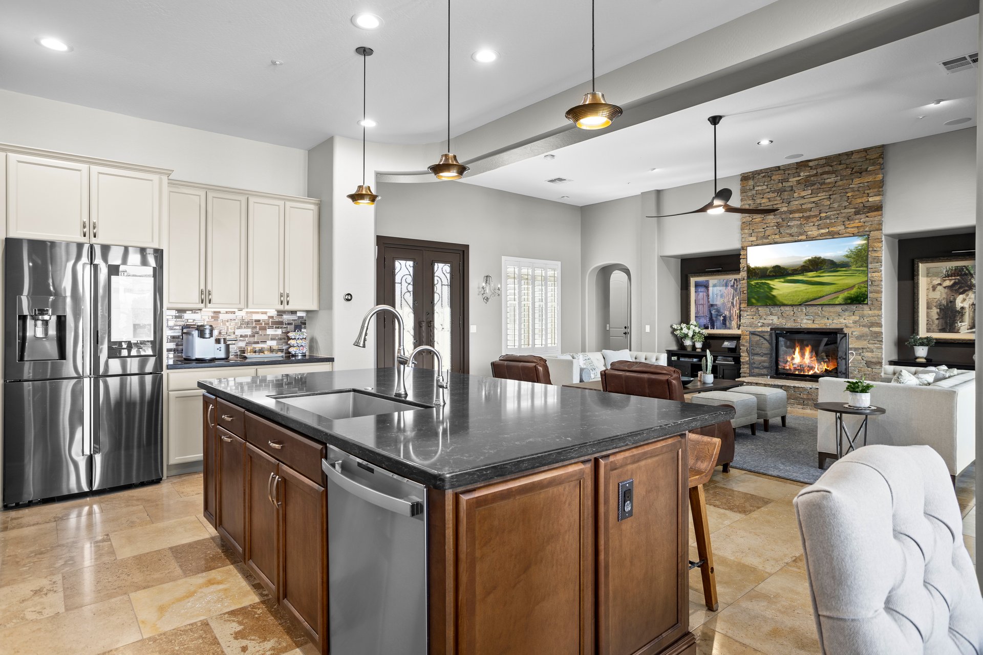 Open-concept kitchen and living room with a large island, stainless steel appliances, pendant lights, and a stone fireplace wall in the background.