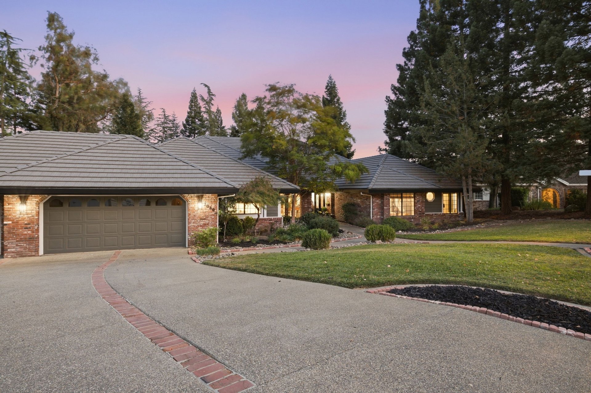 Brick home with a gray tile roof, curved driveway, and illuminated windows at dusk.
