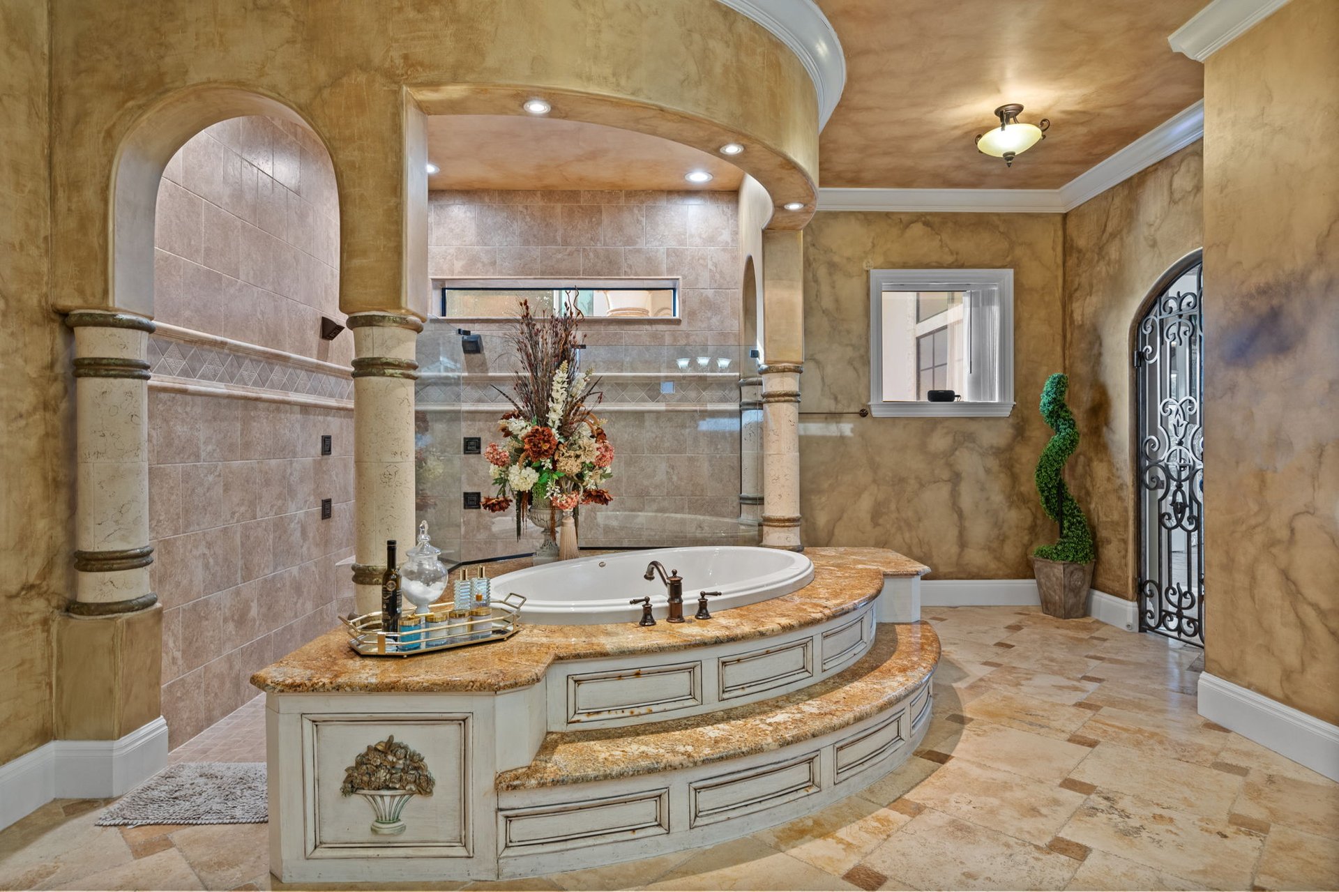 Ornate bathroom with a built-in soaking tub, curved stone vanity, arched openings, and wrought-iron glass door.