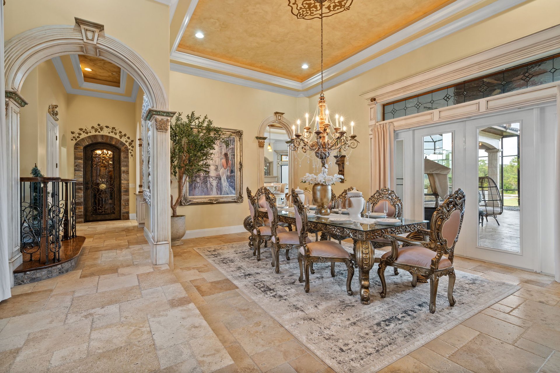 Ornate dining room with a carved table, chandelier, gold-toned ceiling, and glass doors to a patio.
