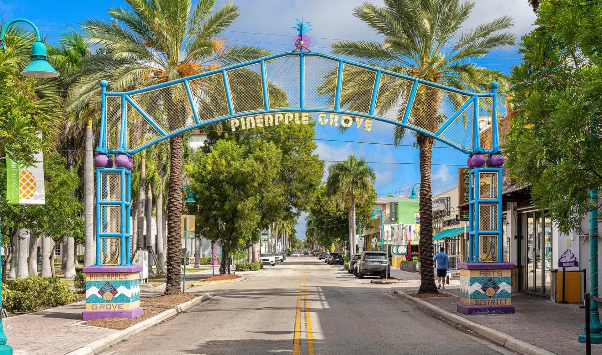 Pineapple Grove Arts District street beneath a blue arch, lined with palm trees and shops.