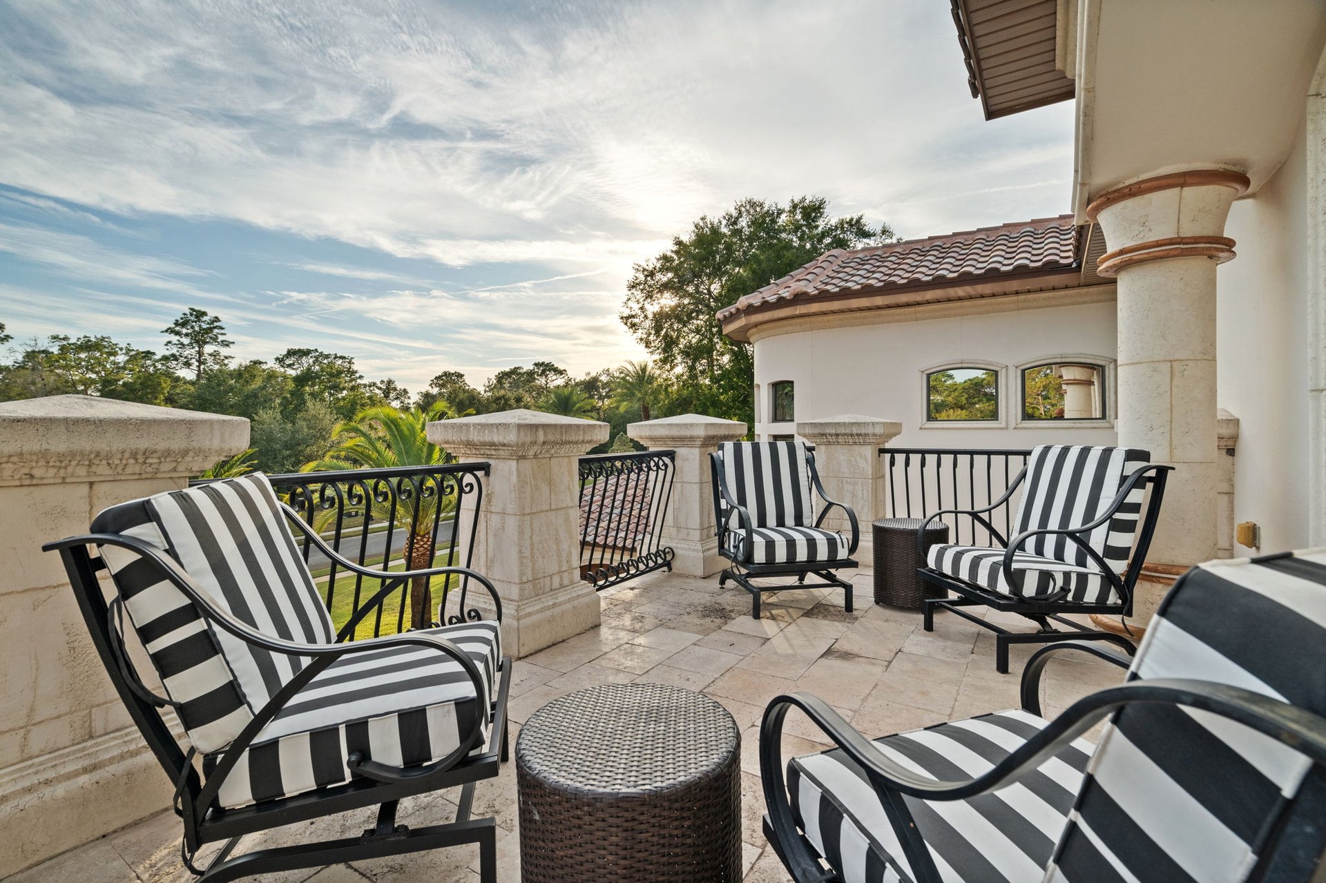 Stone terrace furnished with striped chairs, wrought-iron railings, and views of trees.