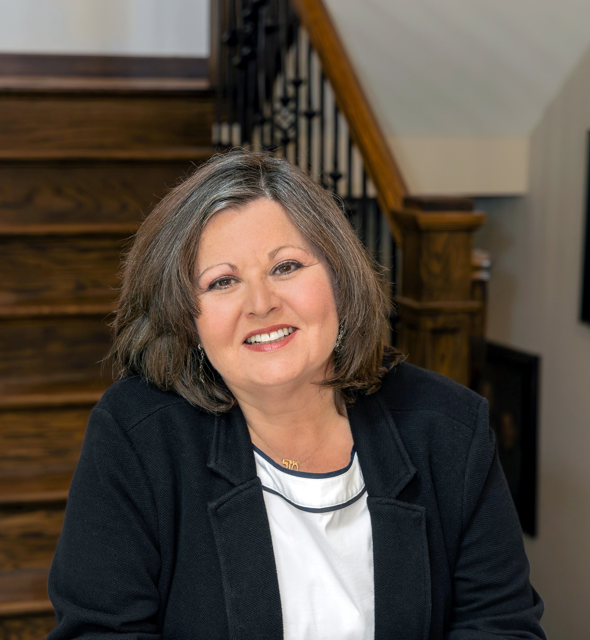 A smiling woman with brown medium-length hair seated in front of a wooden staircase. 