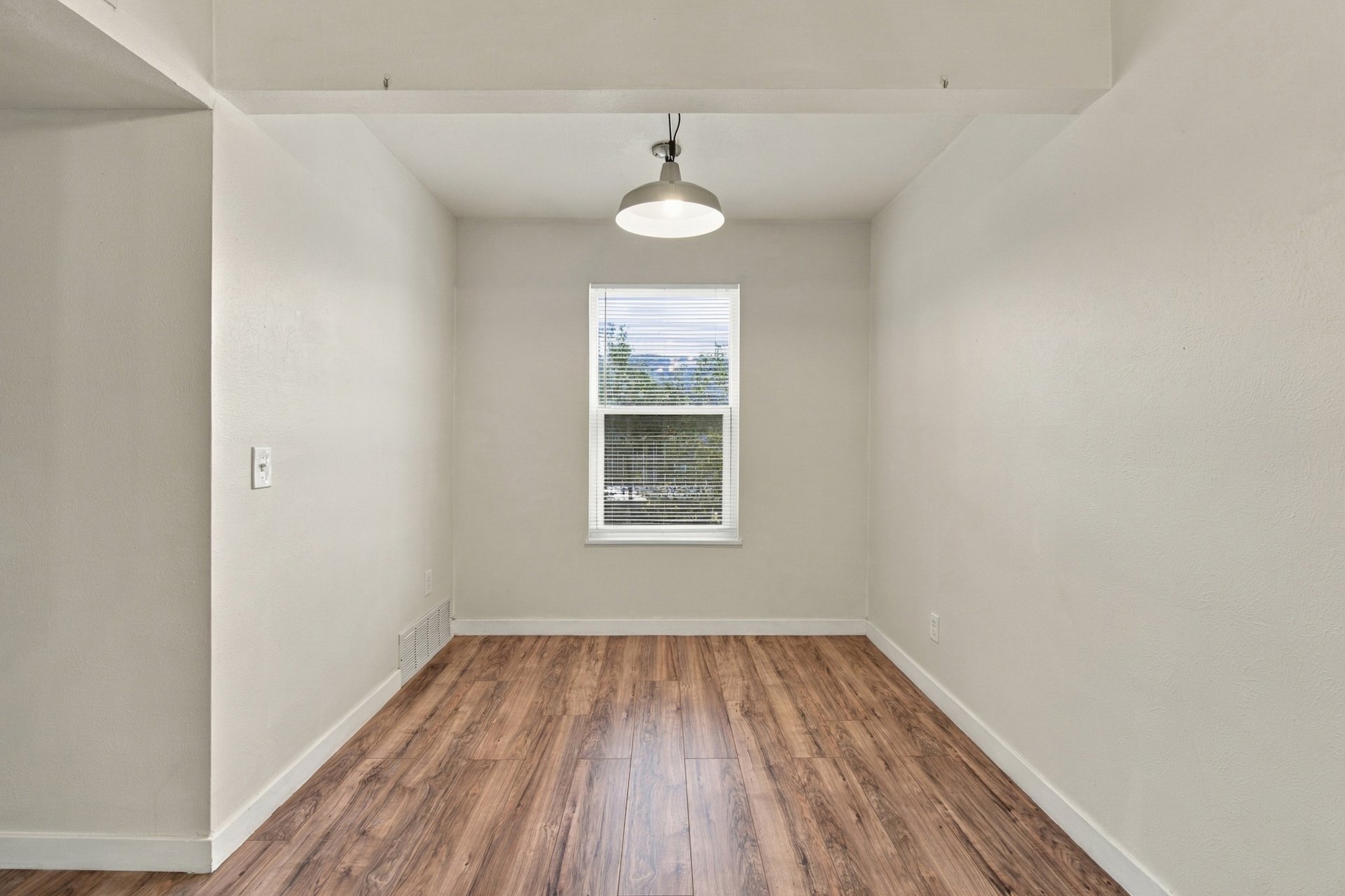 Vacant room with wood-look flooring, a window with blinds, and a hanging ceiling light.