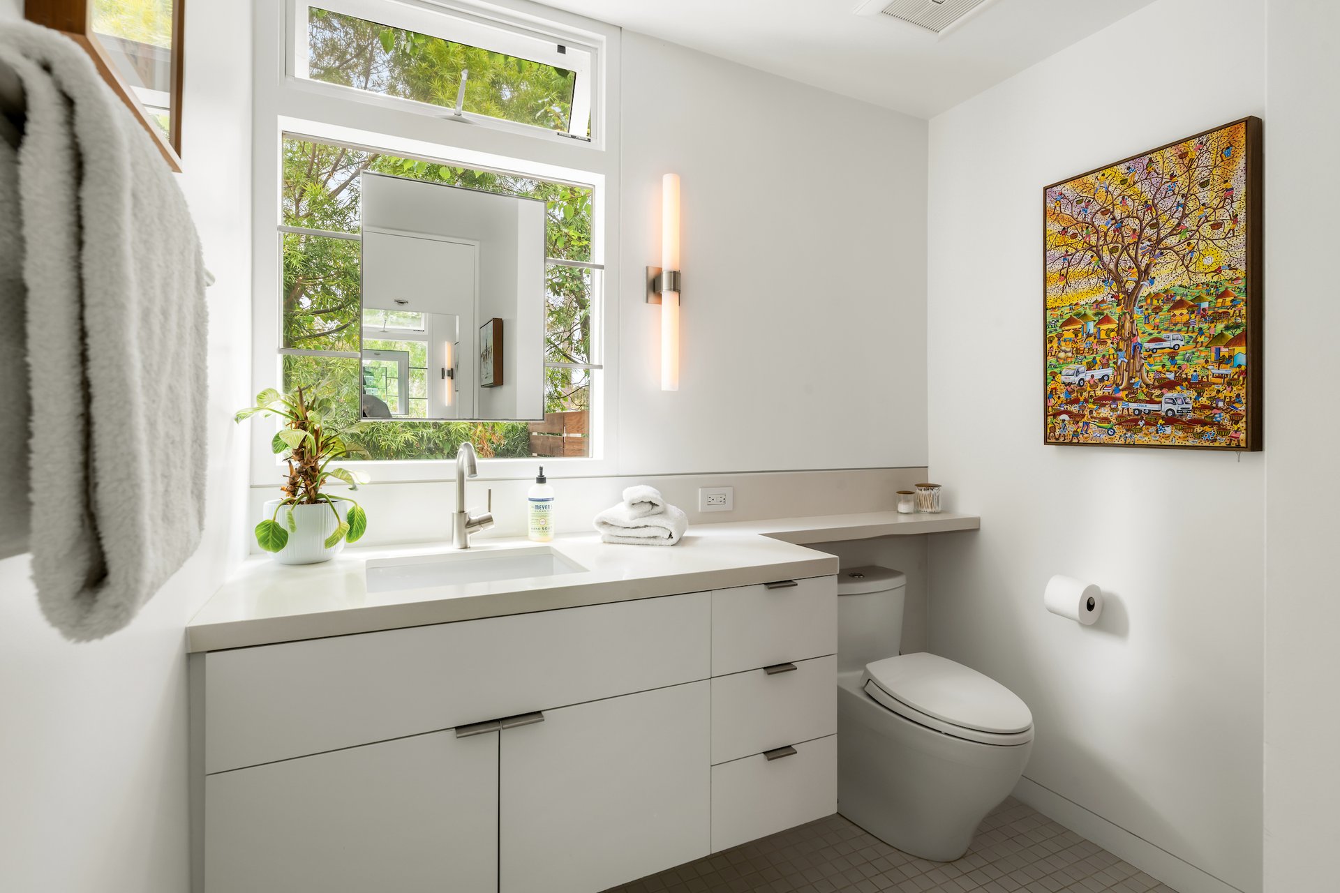 Modern bathroom with a white vanity, large leafy-window view, and colorful framed artwork.