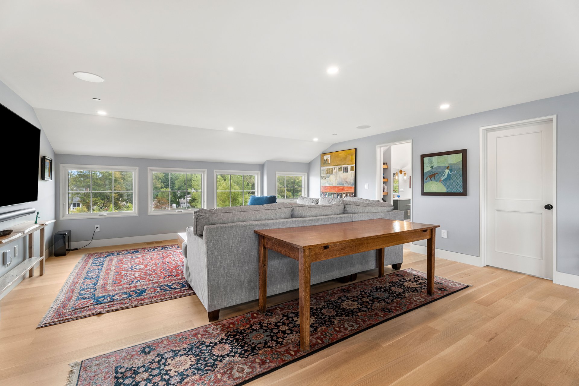 Living room with a gray sectional, broad windows overlooking trees, and patterned rugs on hardwood floors.