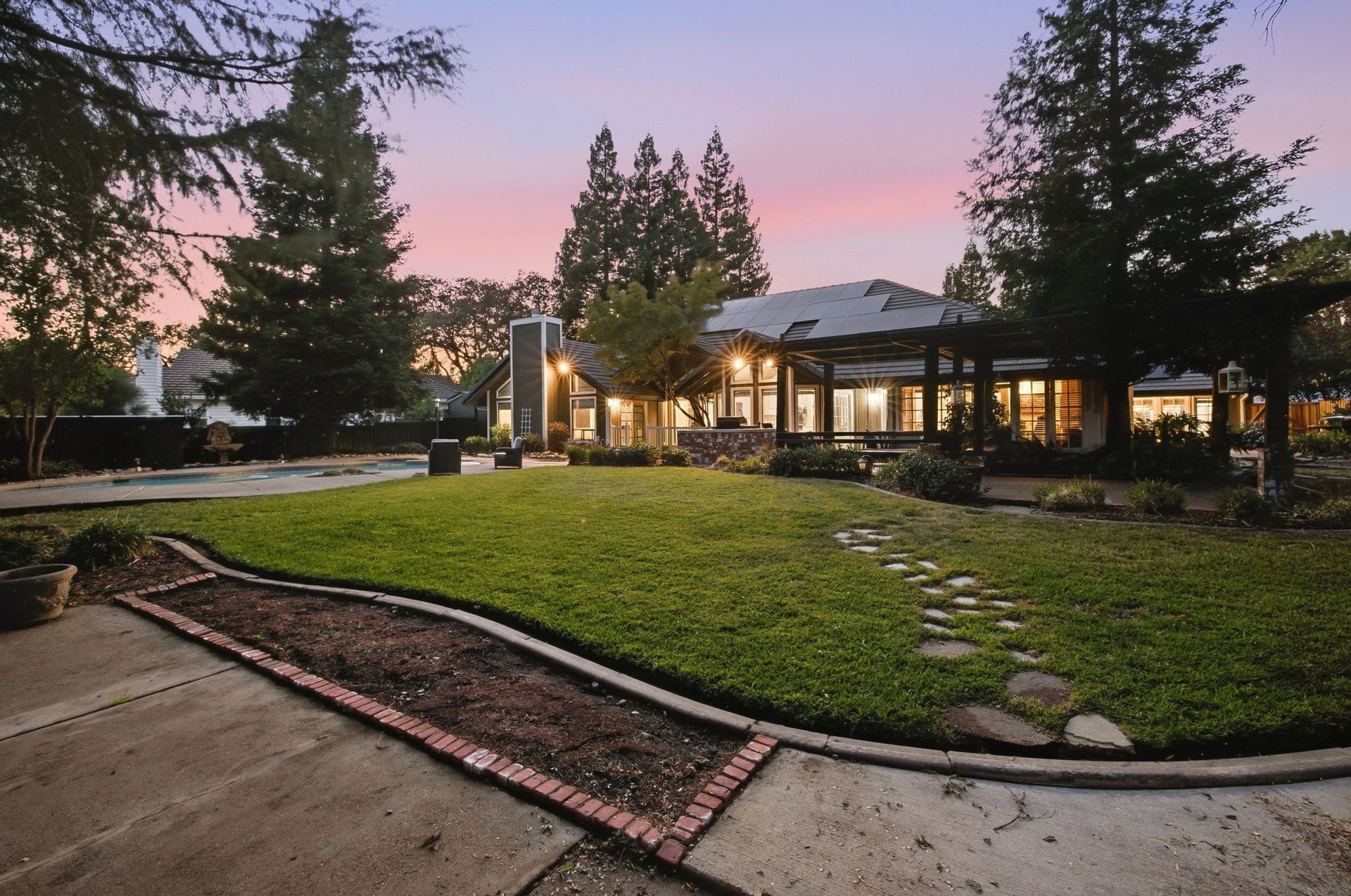 Home’s rear patio and solar-panel roof beside a pool and lawn at dusk