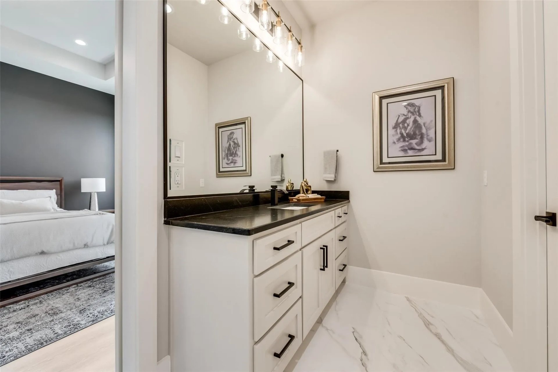 Bathroom vanity with a black countertop and large mirror, with a bedroom visible through the doorway.