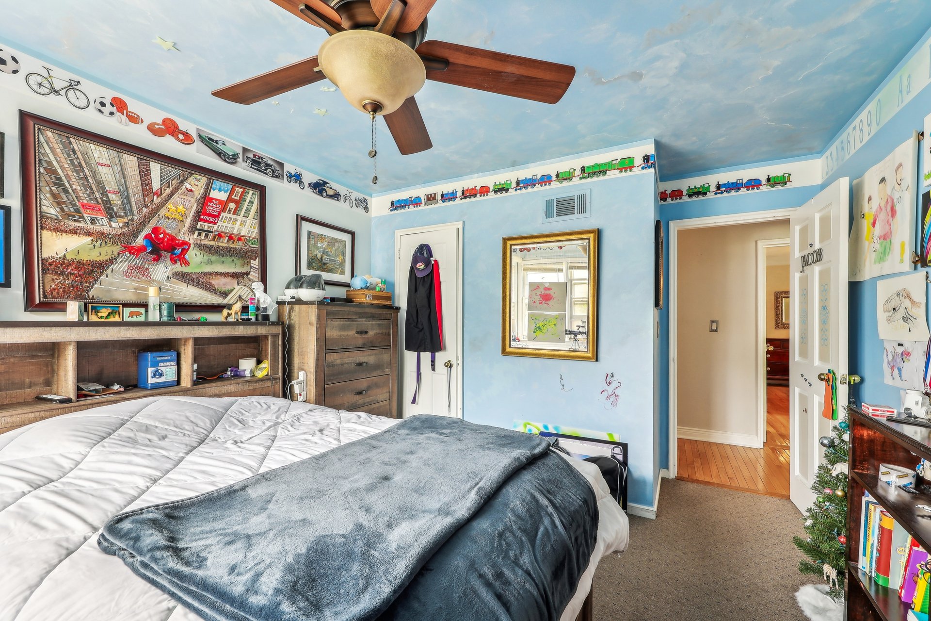 Bedroom with sky-blue walls, a large bed, toy-train borders, and children's drawings.