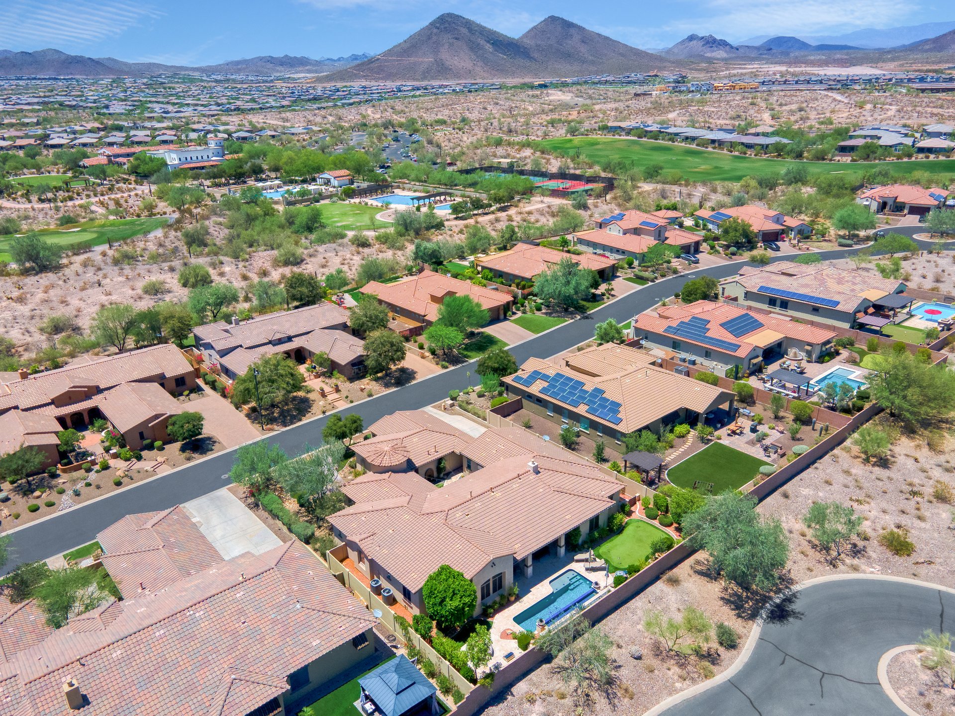 Aerial view of a desert suburban neighborhood with terracotta-roof homes, pools, a golf course, and mountains in the background.
