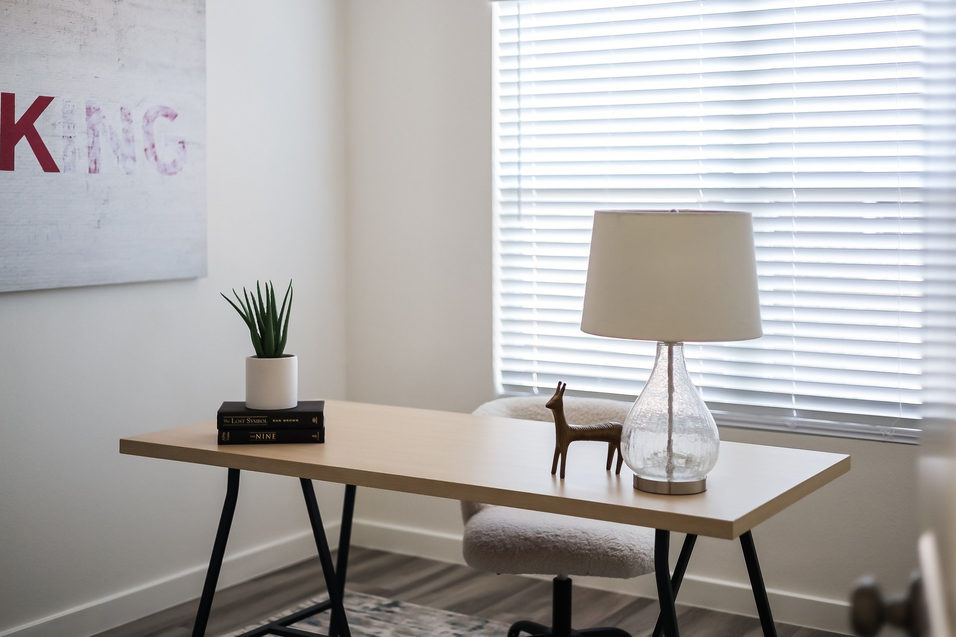 Home office with a light wood desk, glass lamp, potted plant, and bright window with white blinds.
