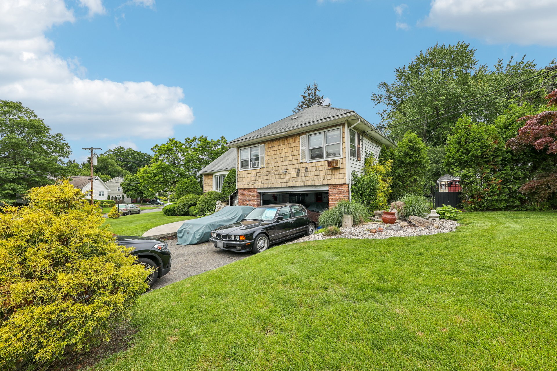 Split-level house with tan shingle siding, an attached garage, driveway cars, and landscaped lawn.