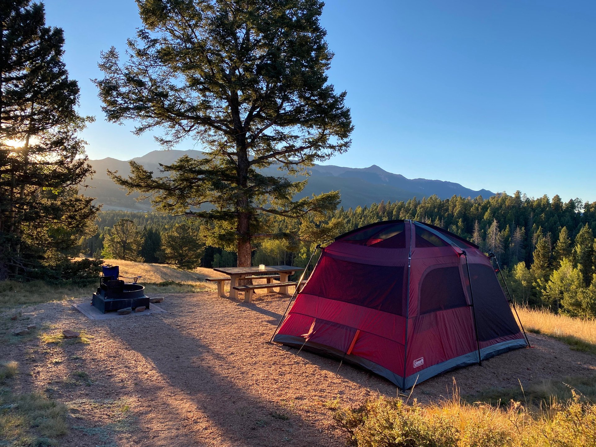 Red tent at a mountain campsite with a picnic table, fire pit, and forested ridges.