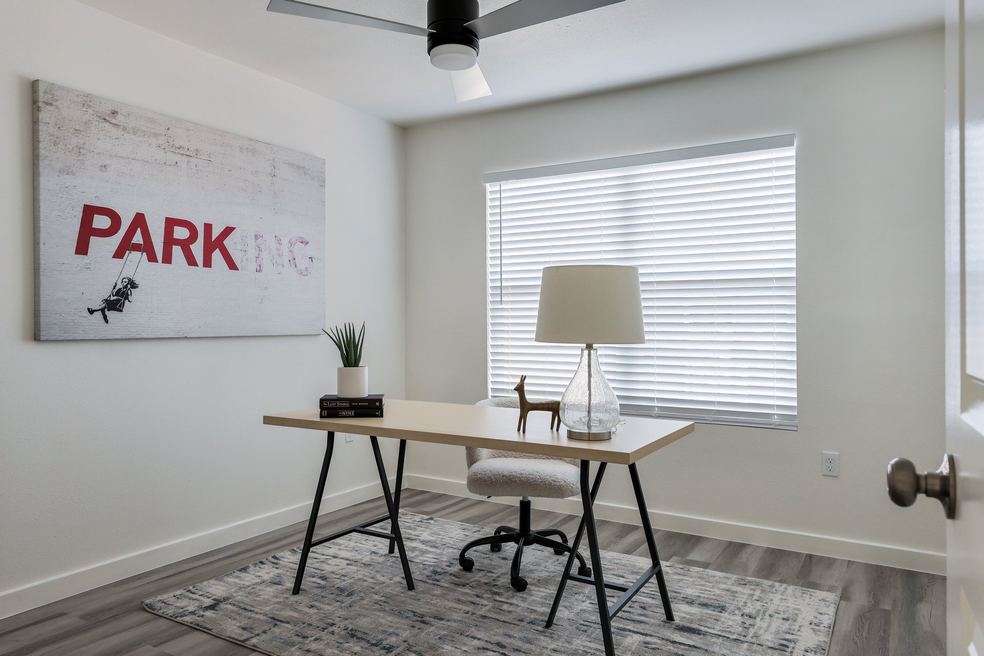 Furnished home office with a light wood desk, patterned rug, and large window with white blinds.