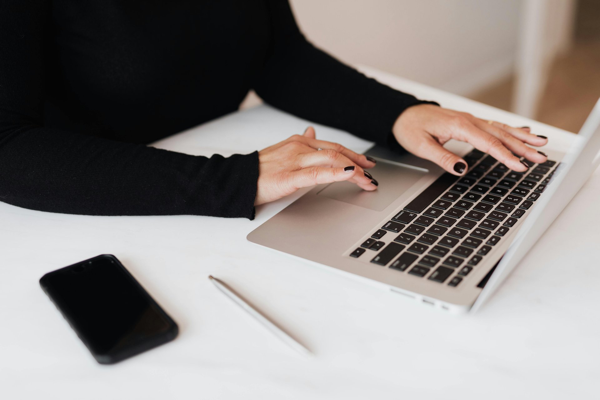 A person typing on a laptop with a smartphone and a pen on a white desk.