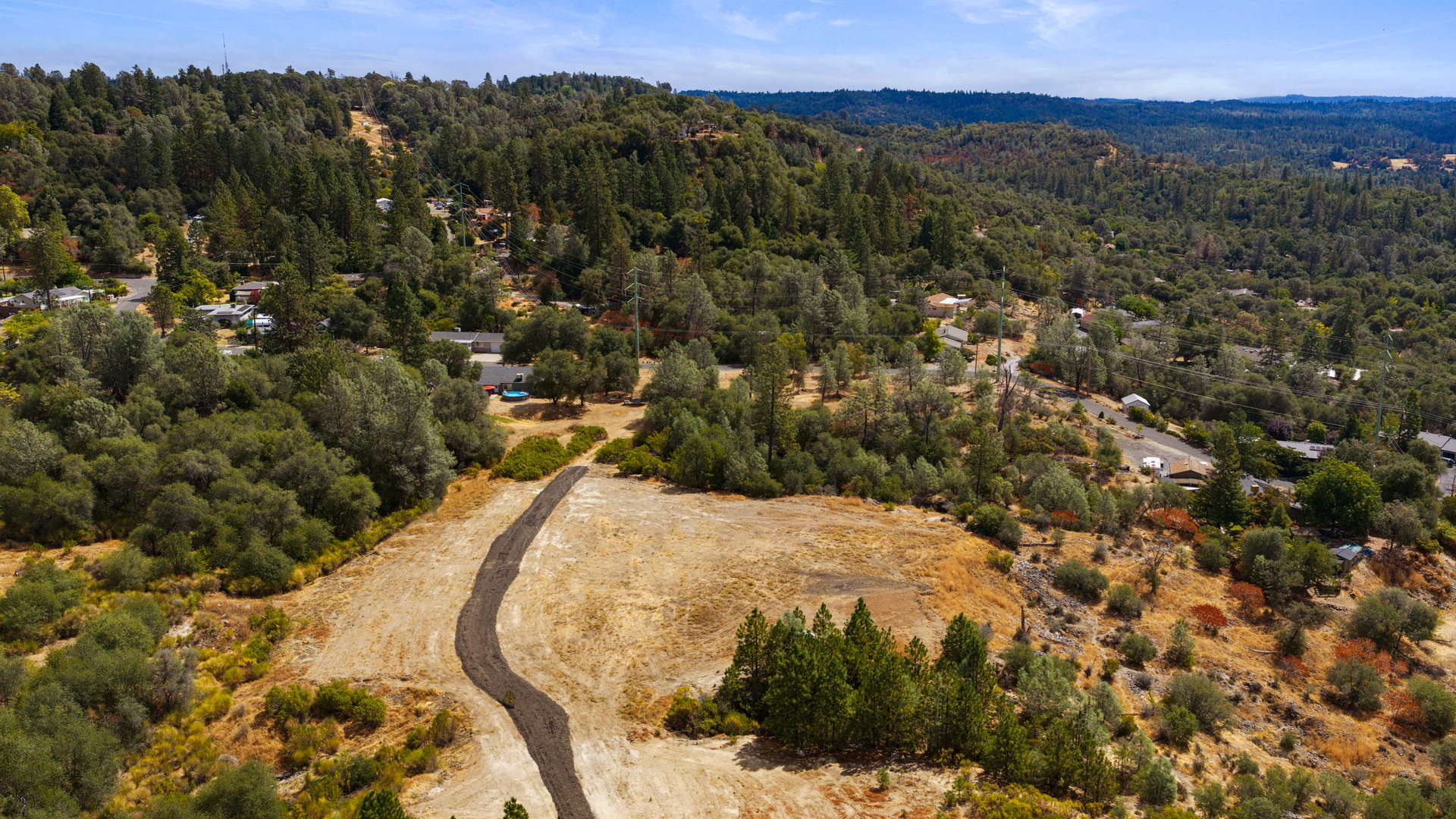 Aerial view of a cleared hillside lot with a winding access road and forested surroundings.