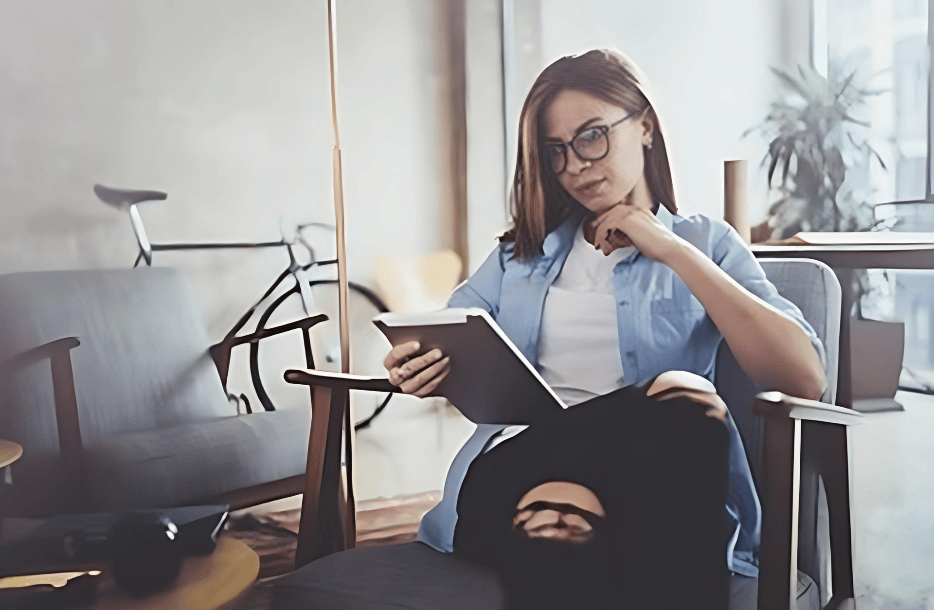 Woman with glasses sits in a modern living room, holding and looking at a tablet.