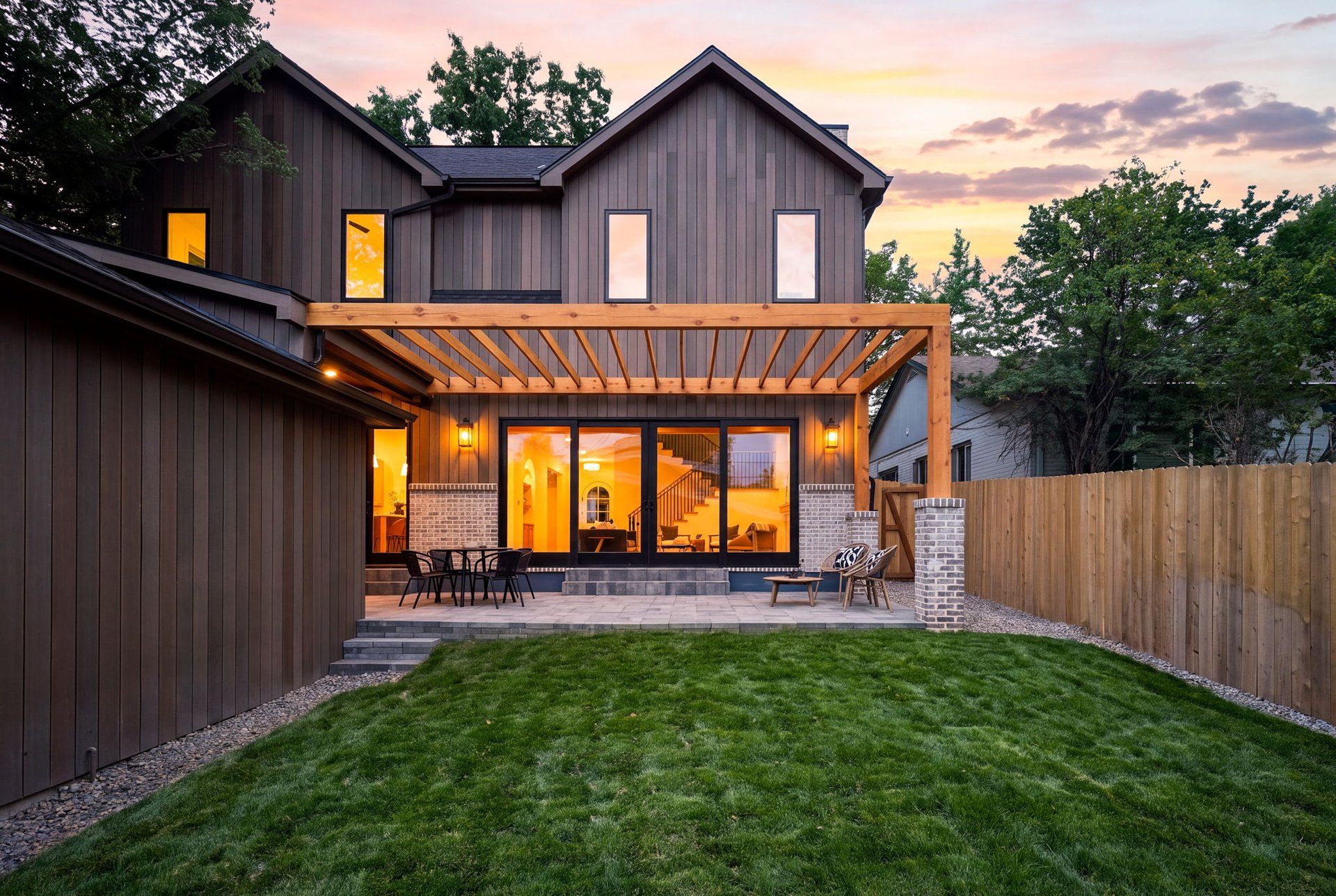 Modern two-story home with a pergola-covered patio, outdoor seating, and a fenced lawn at dusk.