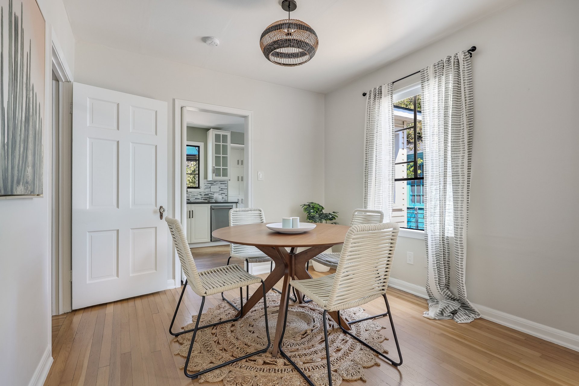 Dining room with a round wood table, woven chairs, and a pendant light beside a curtained window.