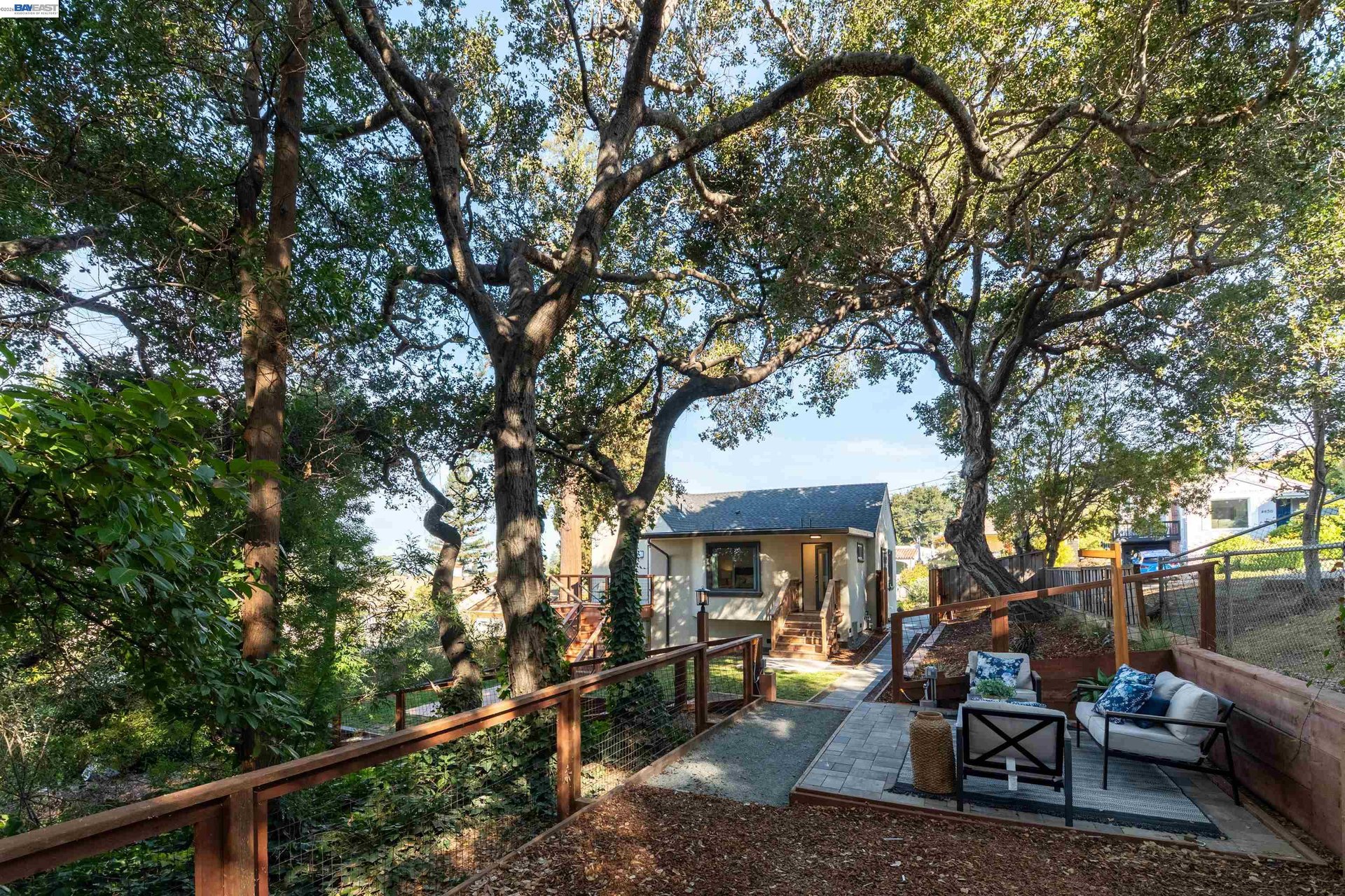 A small house sits beyond a paved patio with outdoor seating beneath mature trees.