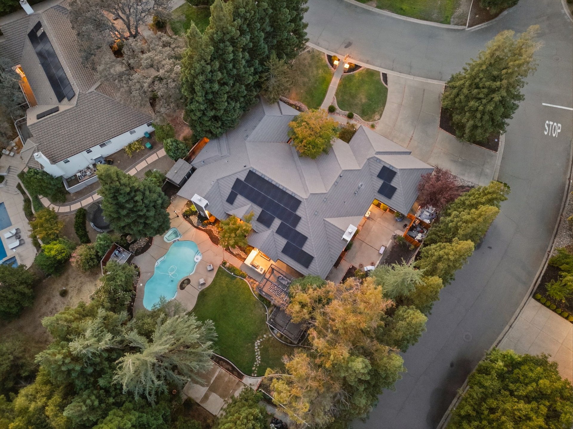 Aerial view of a home with rooftop solar panels, a curved pool, and mature trees.